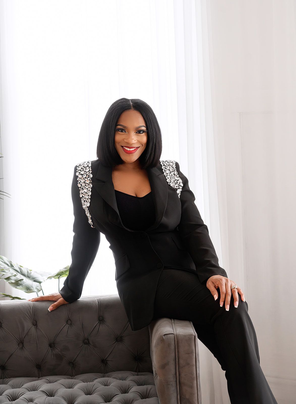 Woman in black sitting on a tufted sofa in a bright room, smiling at the camera.