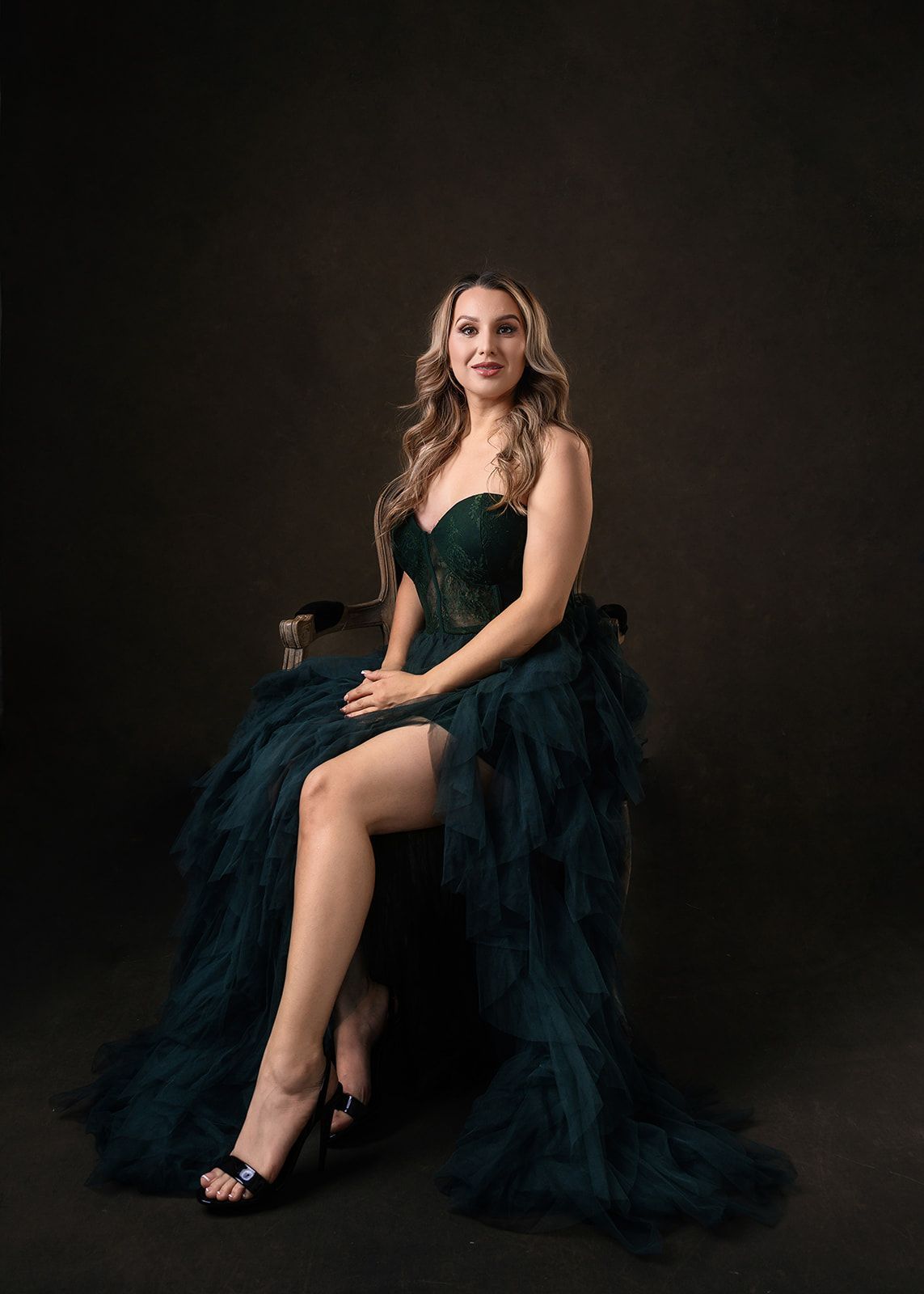 Woman in a dark green gown seated on a stool against a black background.