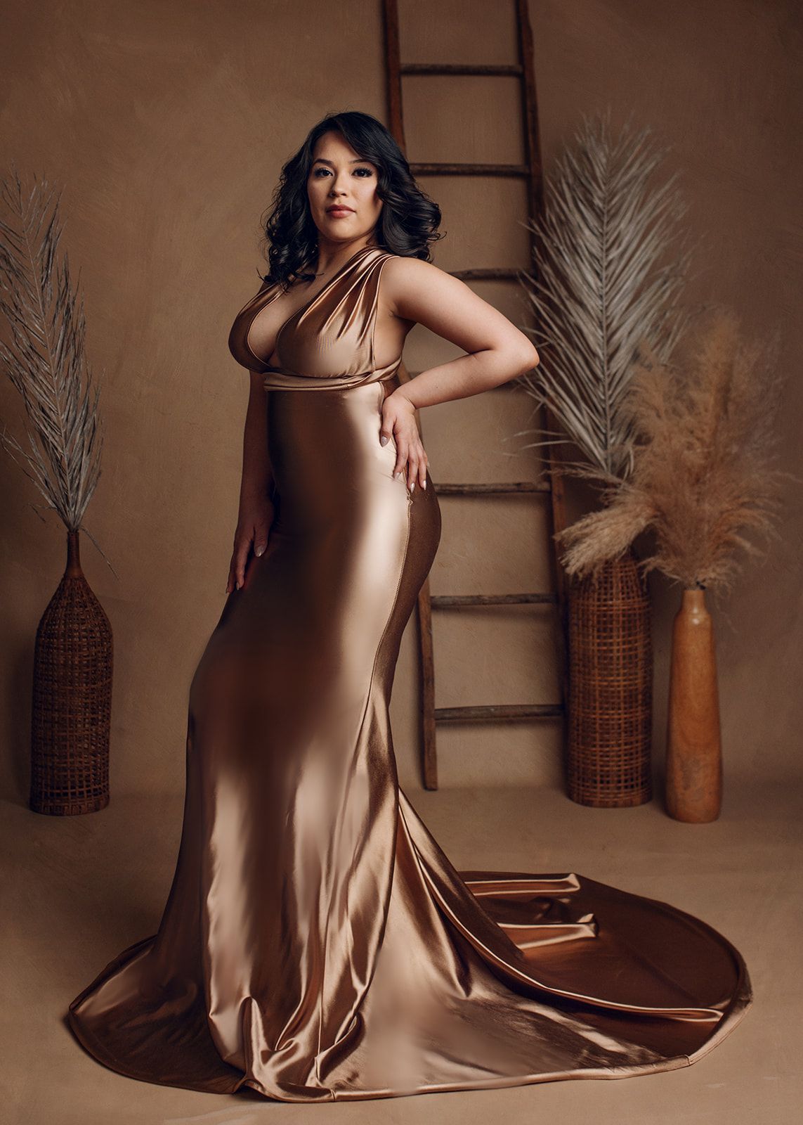 Woman in a bronze satin gown posing beside a ladder and dried grasses in a warm studio setting.
