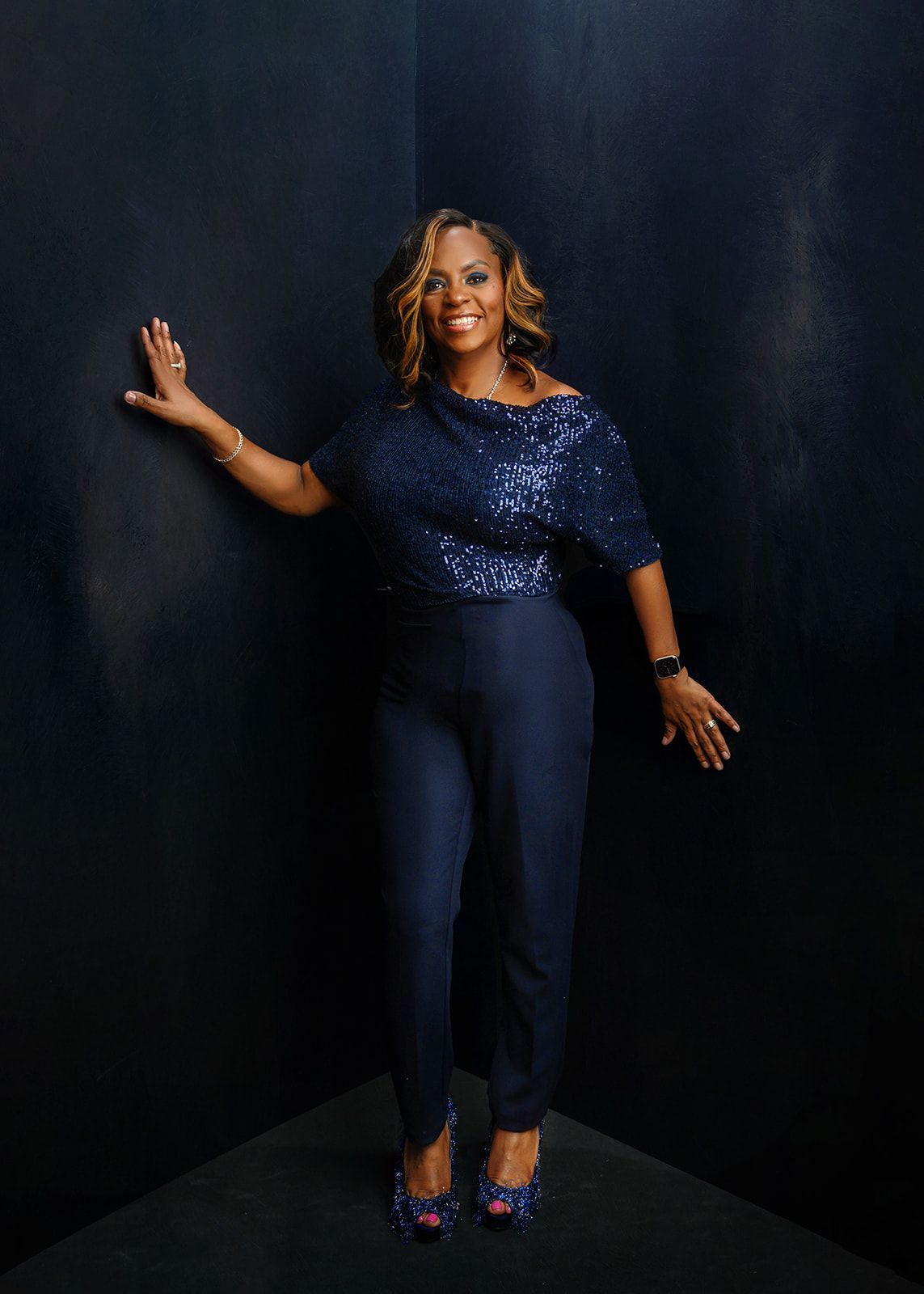 Woman in a navy sequined top and dark pants posing against a dark studio backdrop.