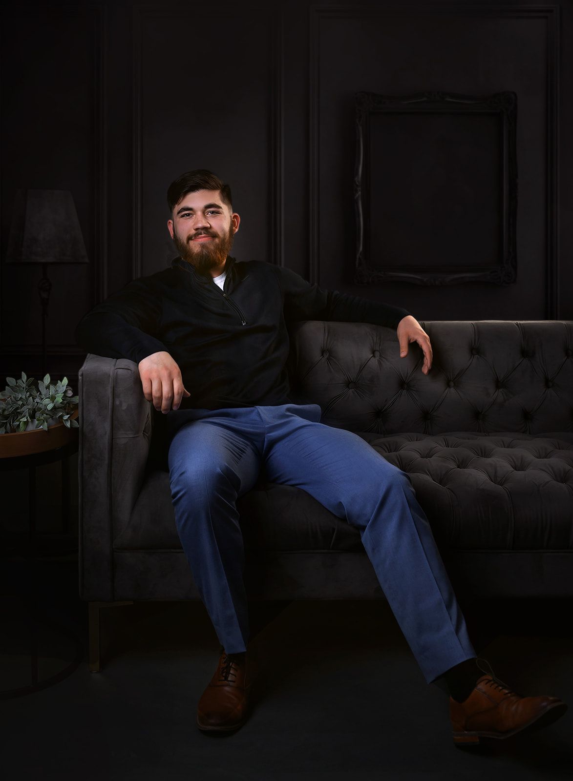 Man seated on a dark tufted sofa in a dim room, wearing dark clothes and jeans, with one arm resting on the backrest.