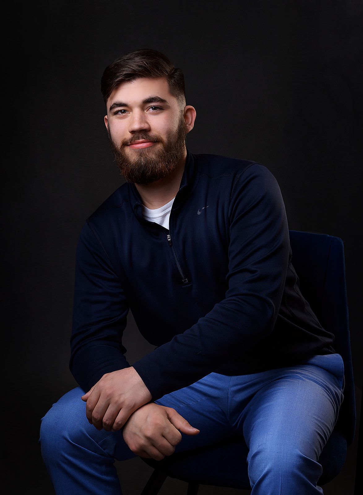 Seated person in a dark sweater and jeans against a black background, smiling softly.