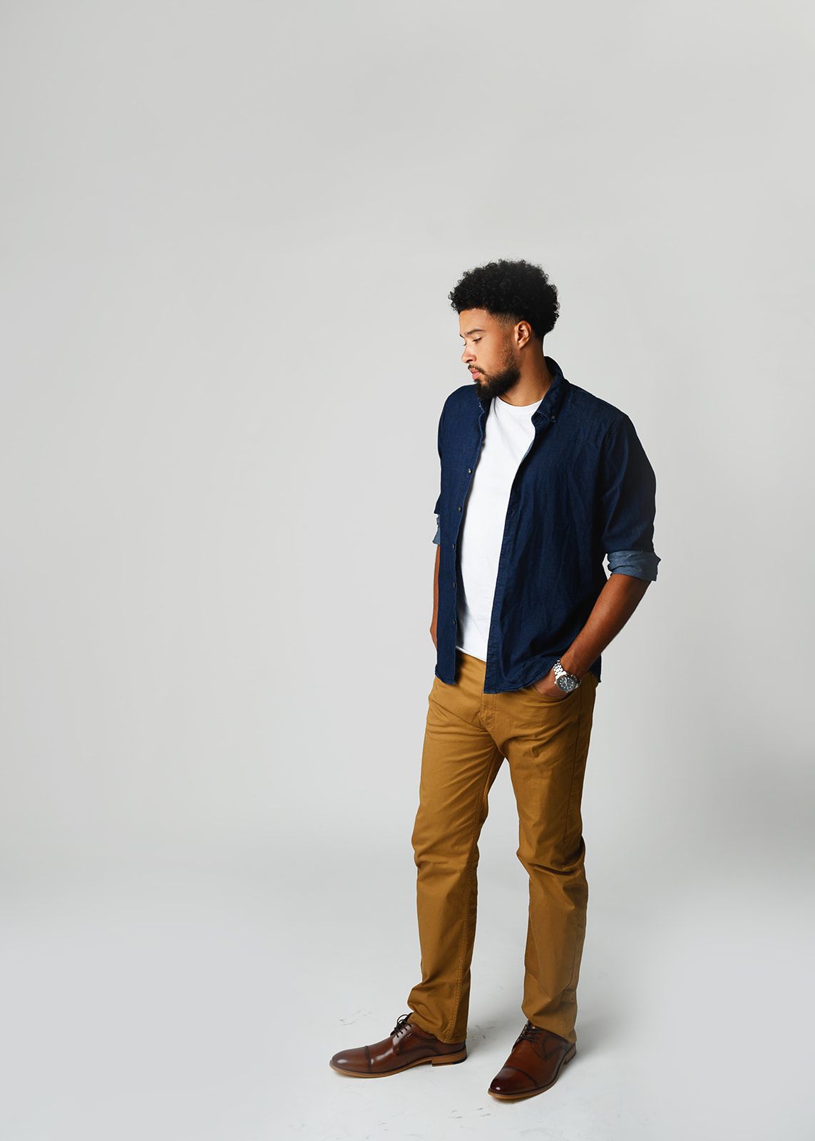 Man standing in a navy shirt, white tee, tan pants, and brown shoes against a light gray background.