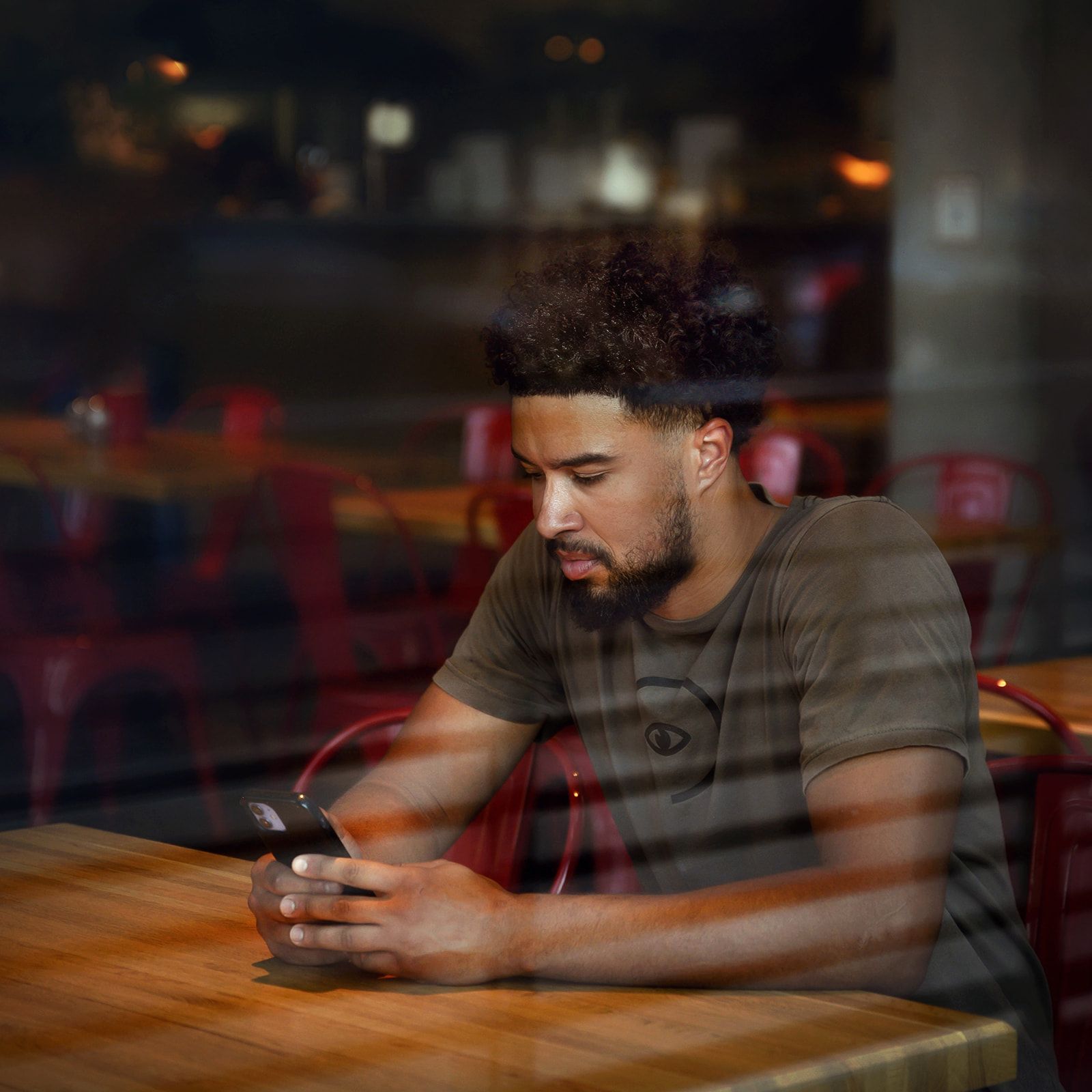 Man seated at a café table, looking at a phone in a dimly lit window reflection.