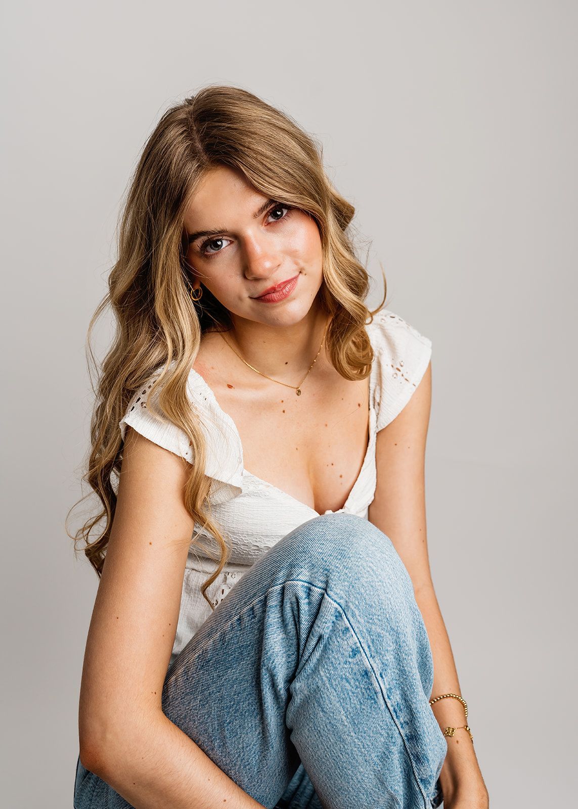 Portrait of a seated woman in a white top and blue jeans, smiling against a plain background.