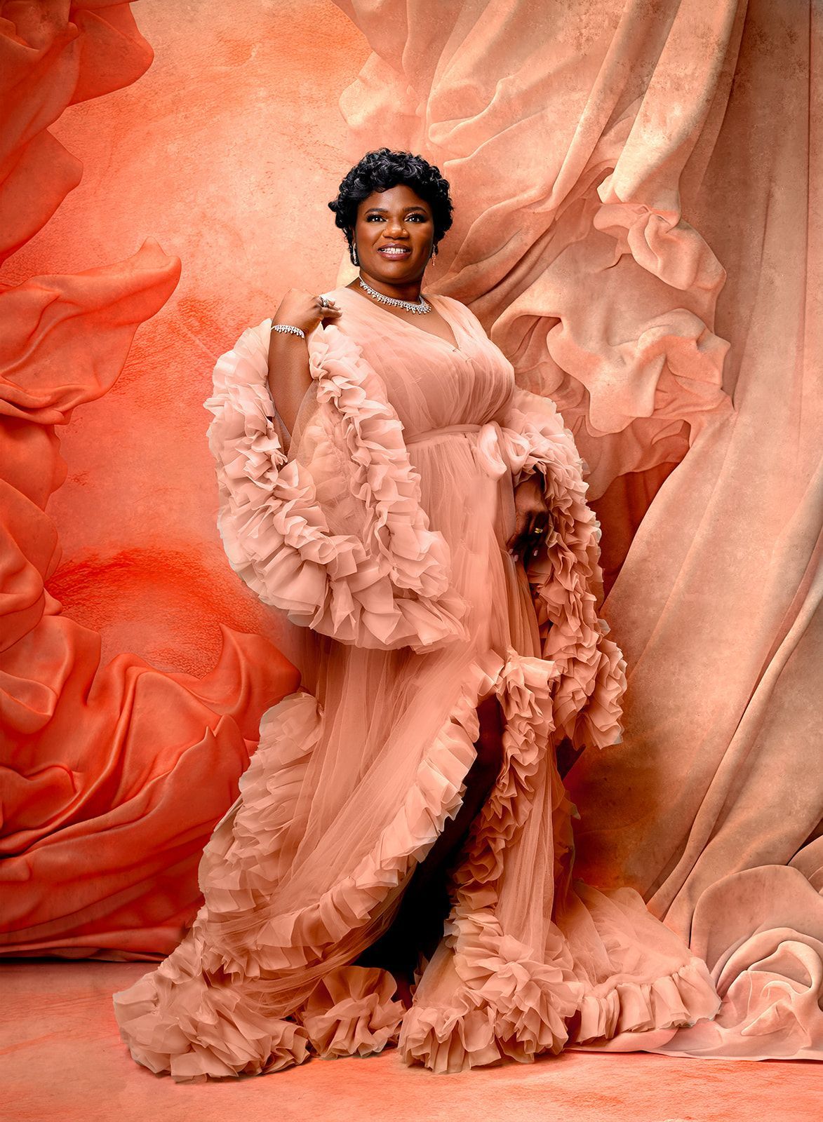 Person in a blush ruffled gown posing against flowing coral and peach fabric backdrop.
