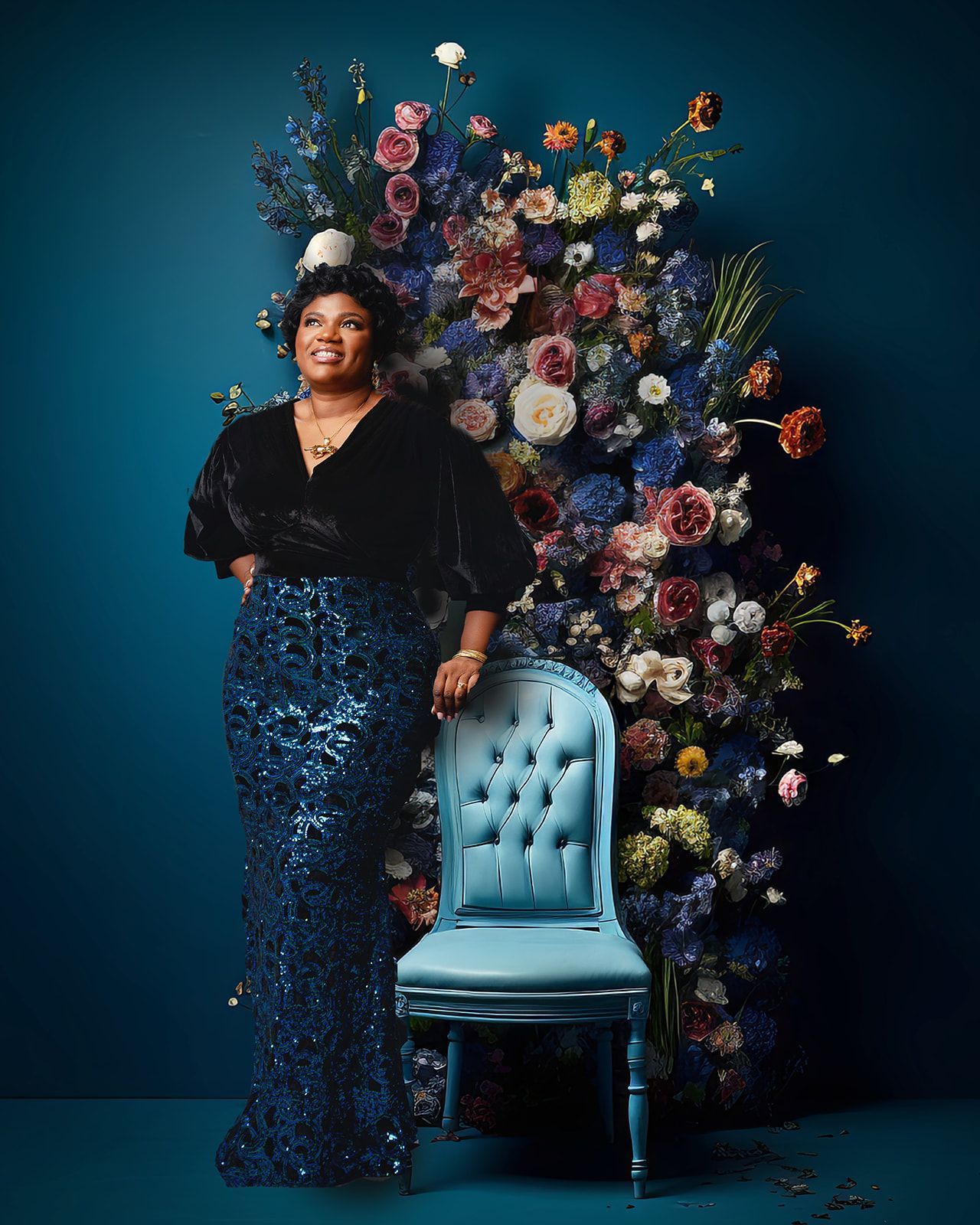 Woman in a sequined navy gown standing beside a blue tufted chair and a floral backdrop.