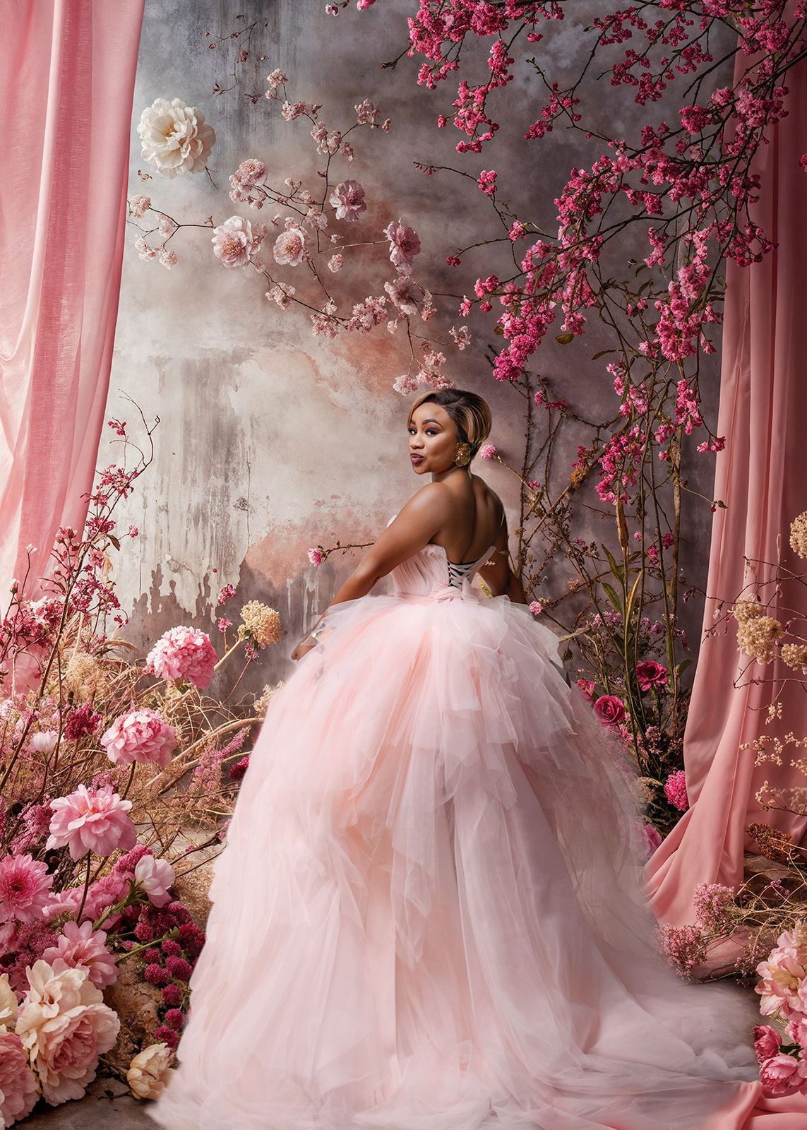Woman in a pink tulle gown standing among pink flowers and draped curtains in a floral studio setting.