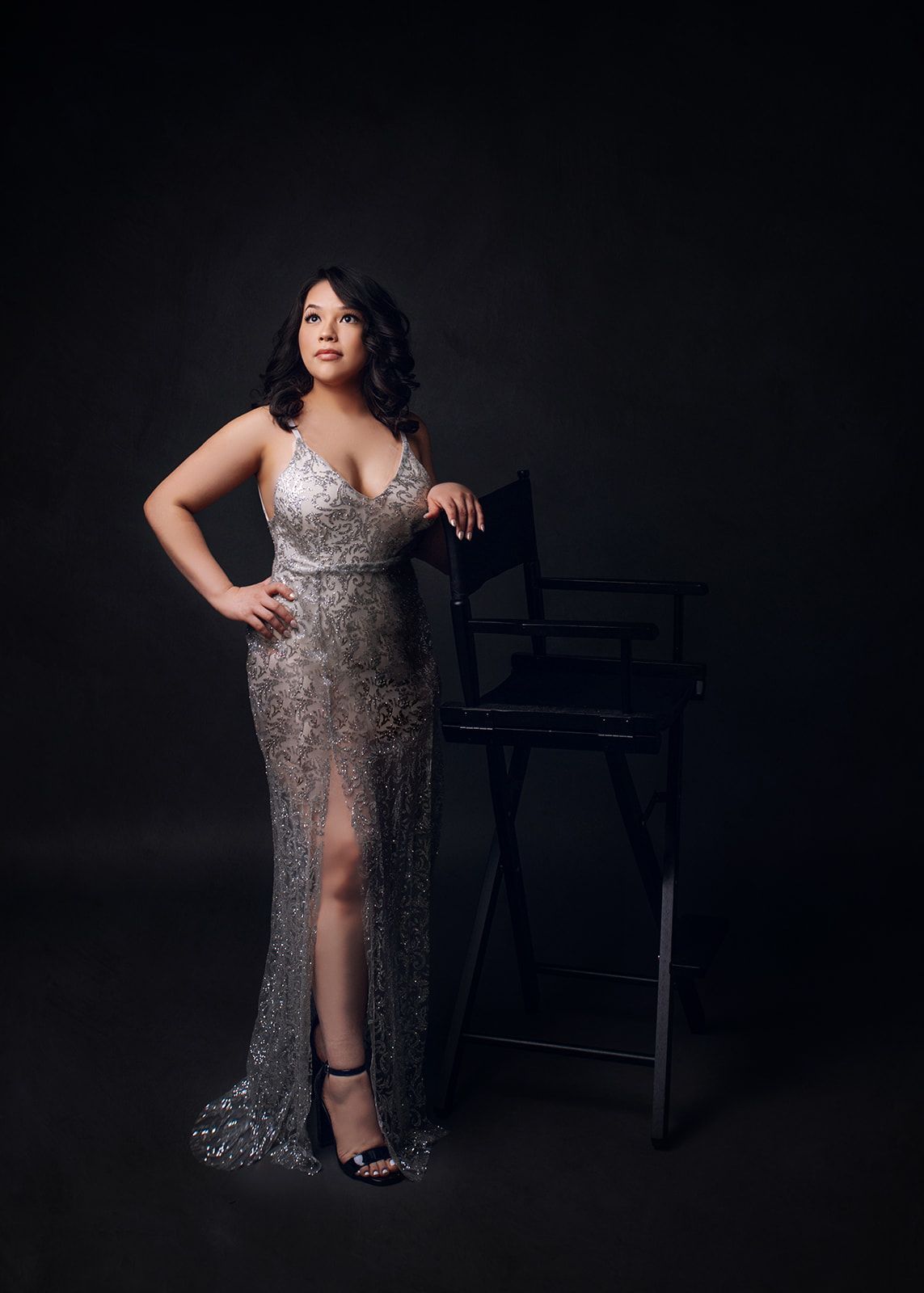 Woman in a silver sequined gown posing beside a high chair against a dark backdrop.