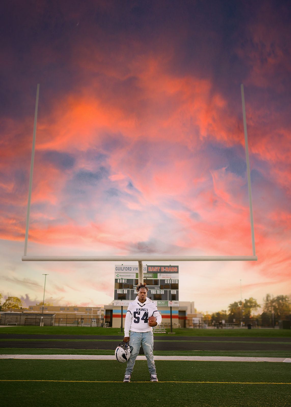 Football player standing on a field at sunset beneath a vivid pink-orange sky.