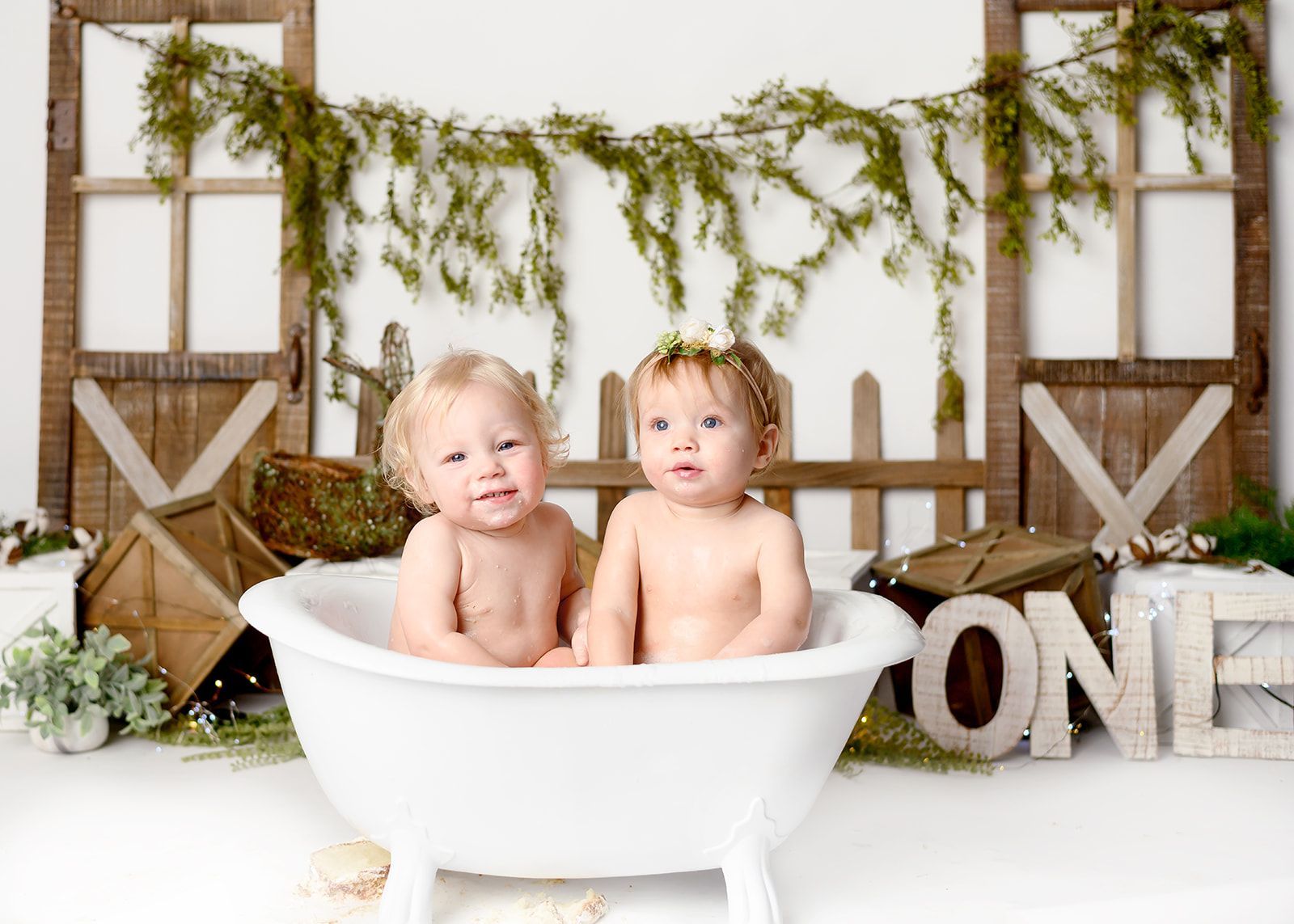 Two babies sitting in a white bathtub, with rustic decor and greenery behind them.