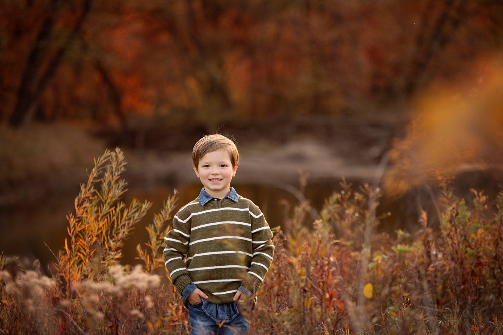 Smiling child in a striped sweater standing in tall autumn grass with warm blurred foliage behind him.