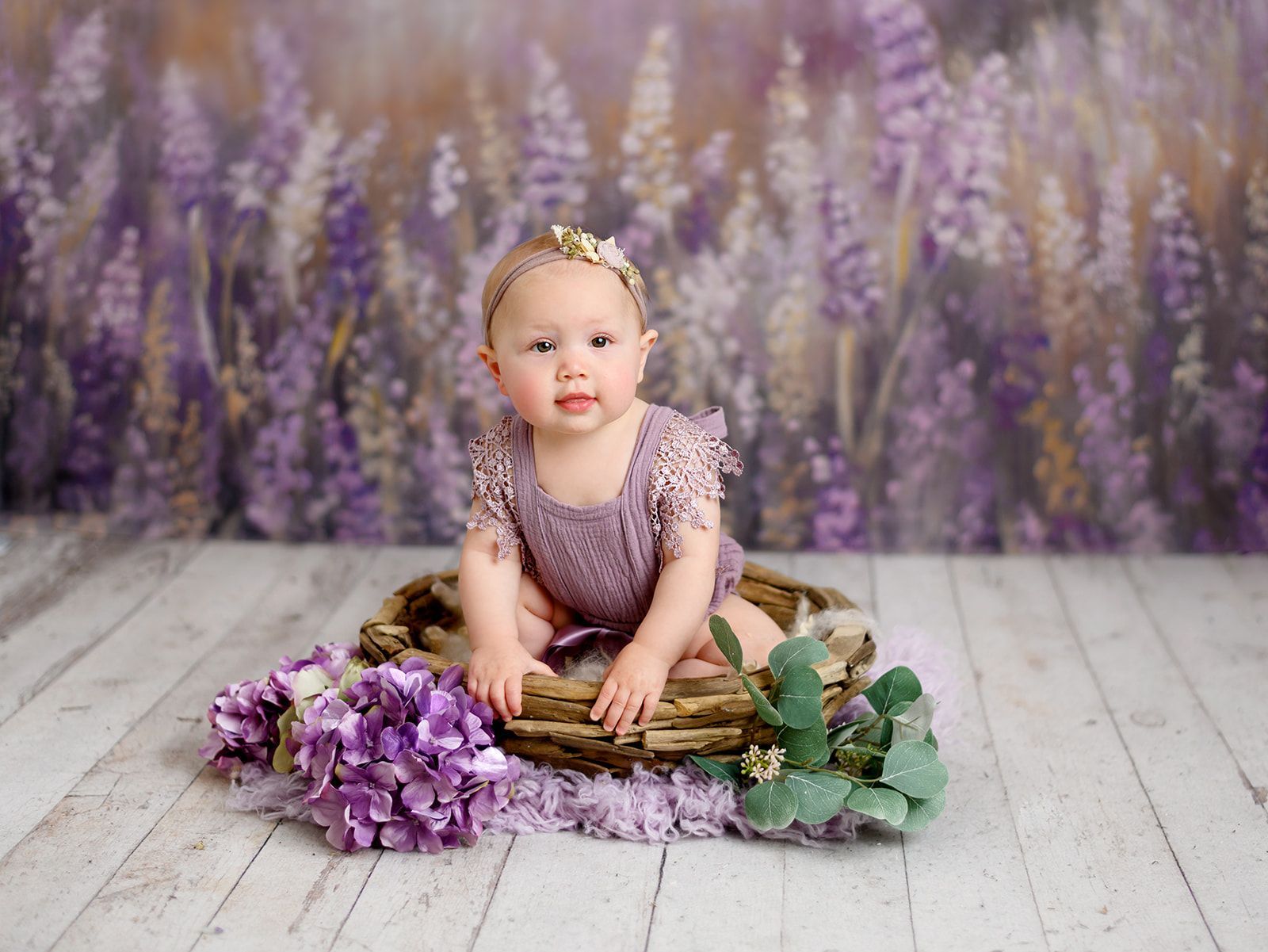 Baby sitting on a wooden basket with purple flowers in a lavender studio backdrop.