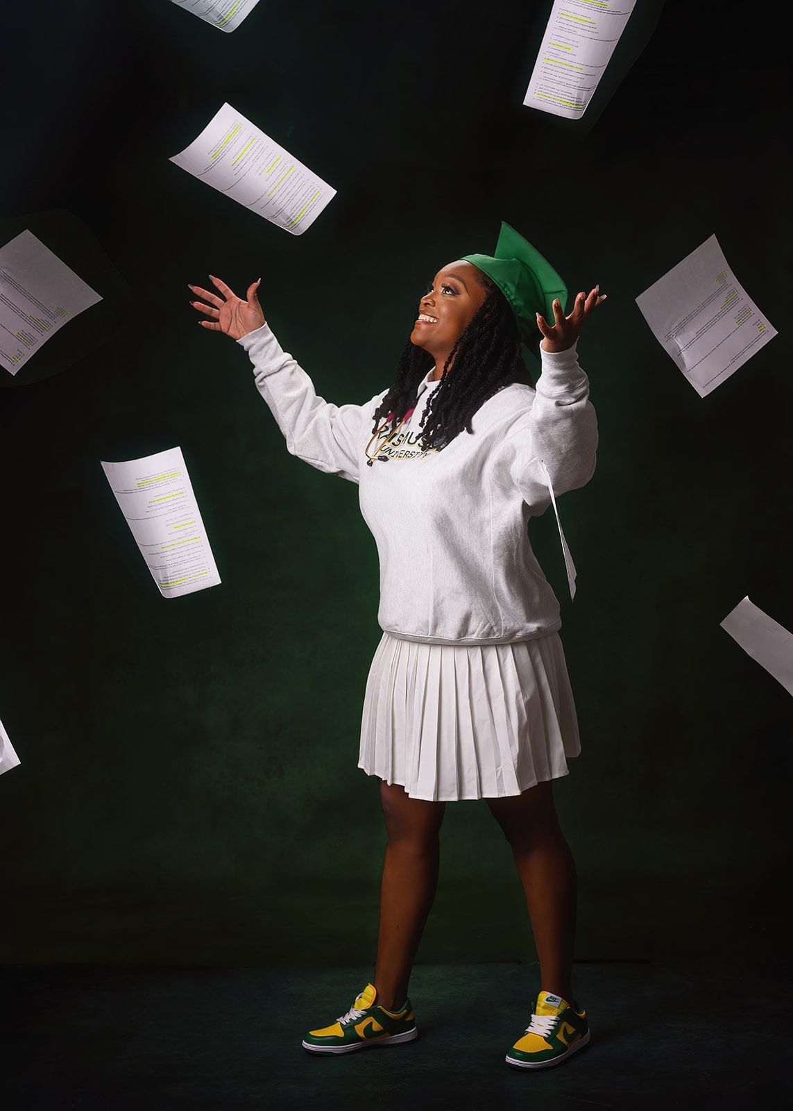 Person in white sweater and pleated skirt with green hat, arms raised as papers float around against a dark background.