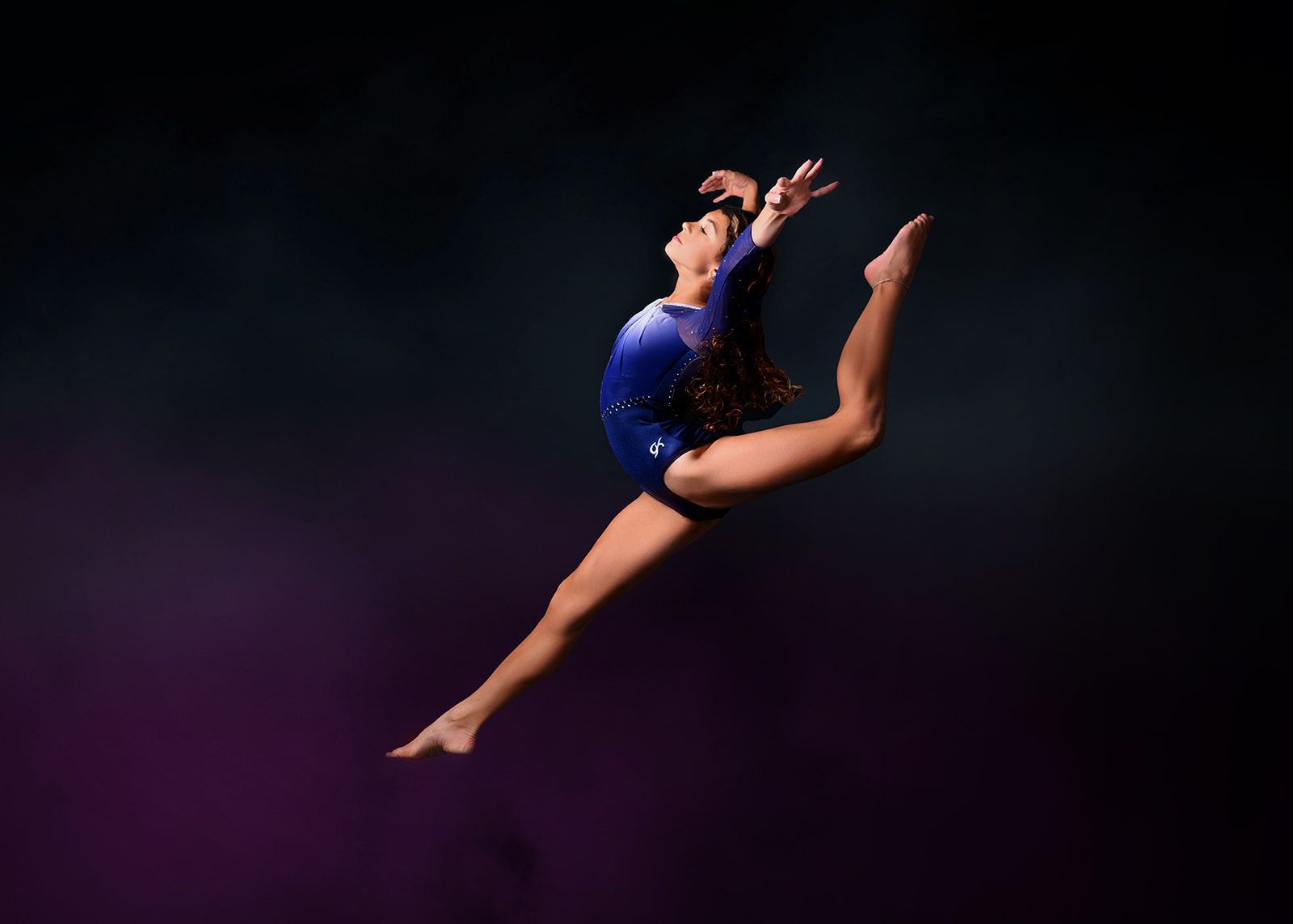 Ballet dancer in a blue costume leaping against a dark purple stage background.