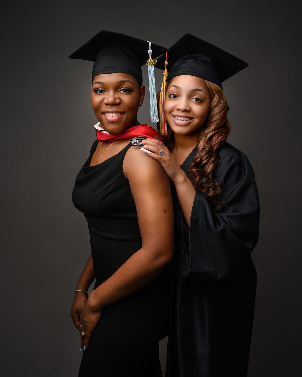 Two graduates in black caps and gowns posing together against a dark backdrop.