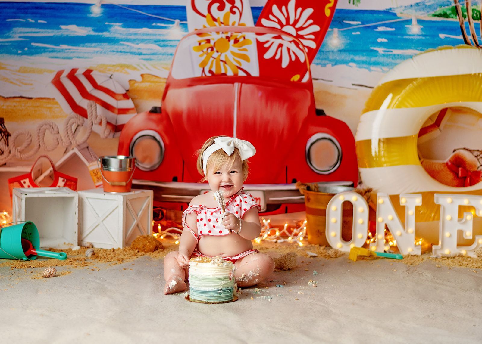 Baby sitting on a beach-themed backdrop with a red truck and large “ONE” letters.