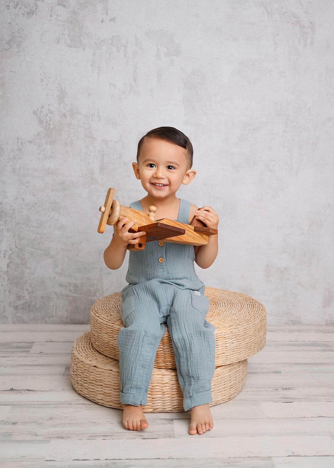 Smiling child sitting on woven cushions, holding a wooden toy airplane against a light gray wall.