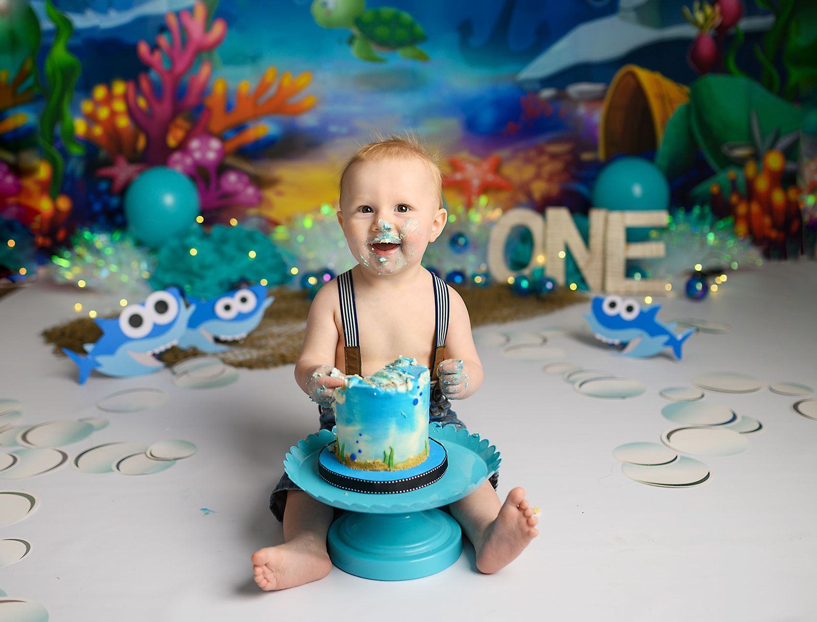 Toddler seated by a blue cake stand, smiling in a colorful underwater-themed birthday setup with fish decorations.