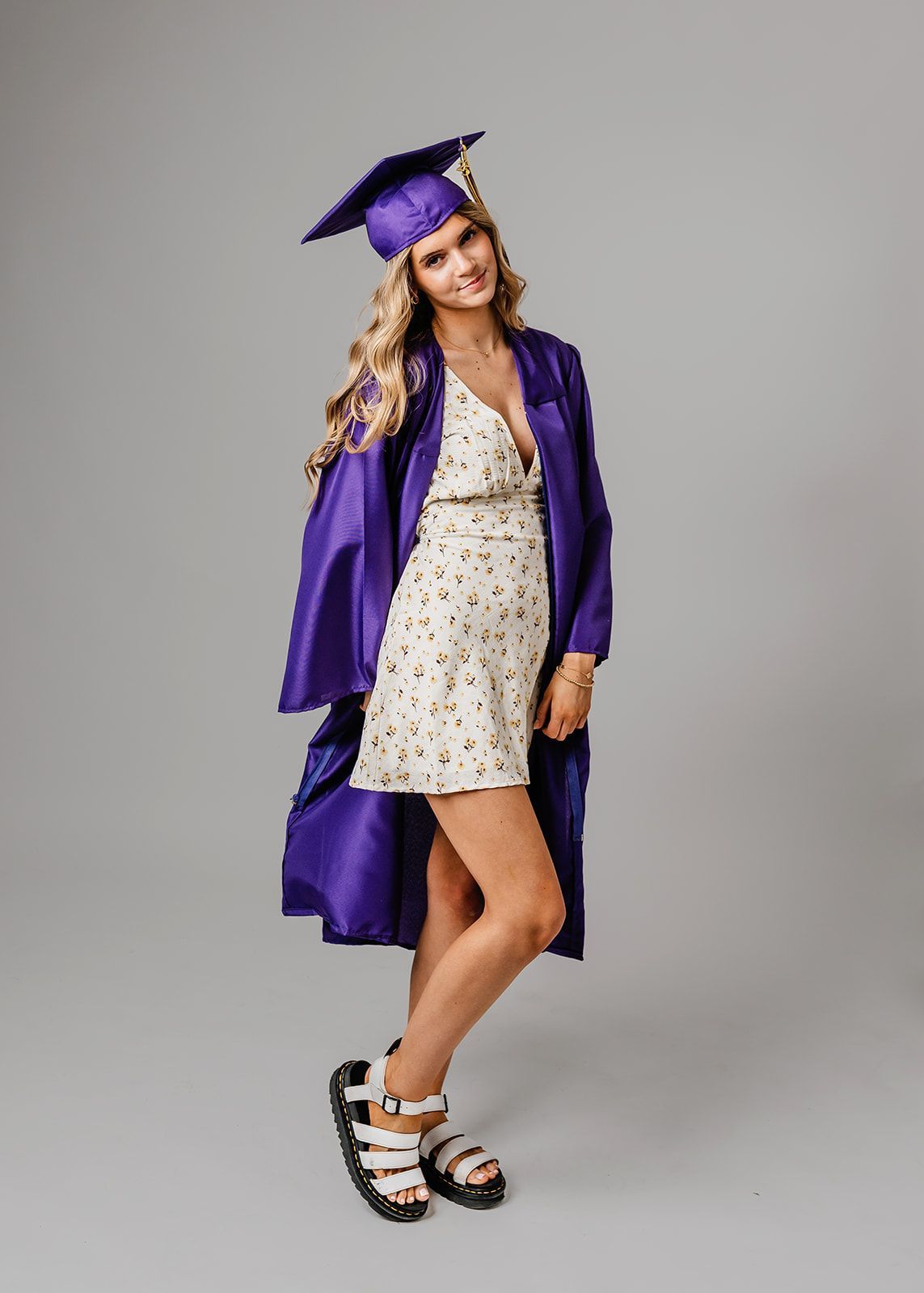 Woman in a purple graduation gown and cap posing in striped platform sandals against a gray backdrop.