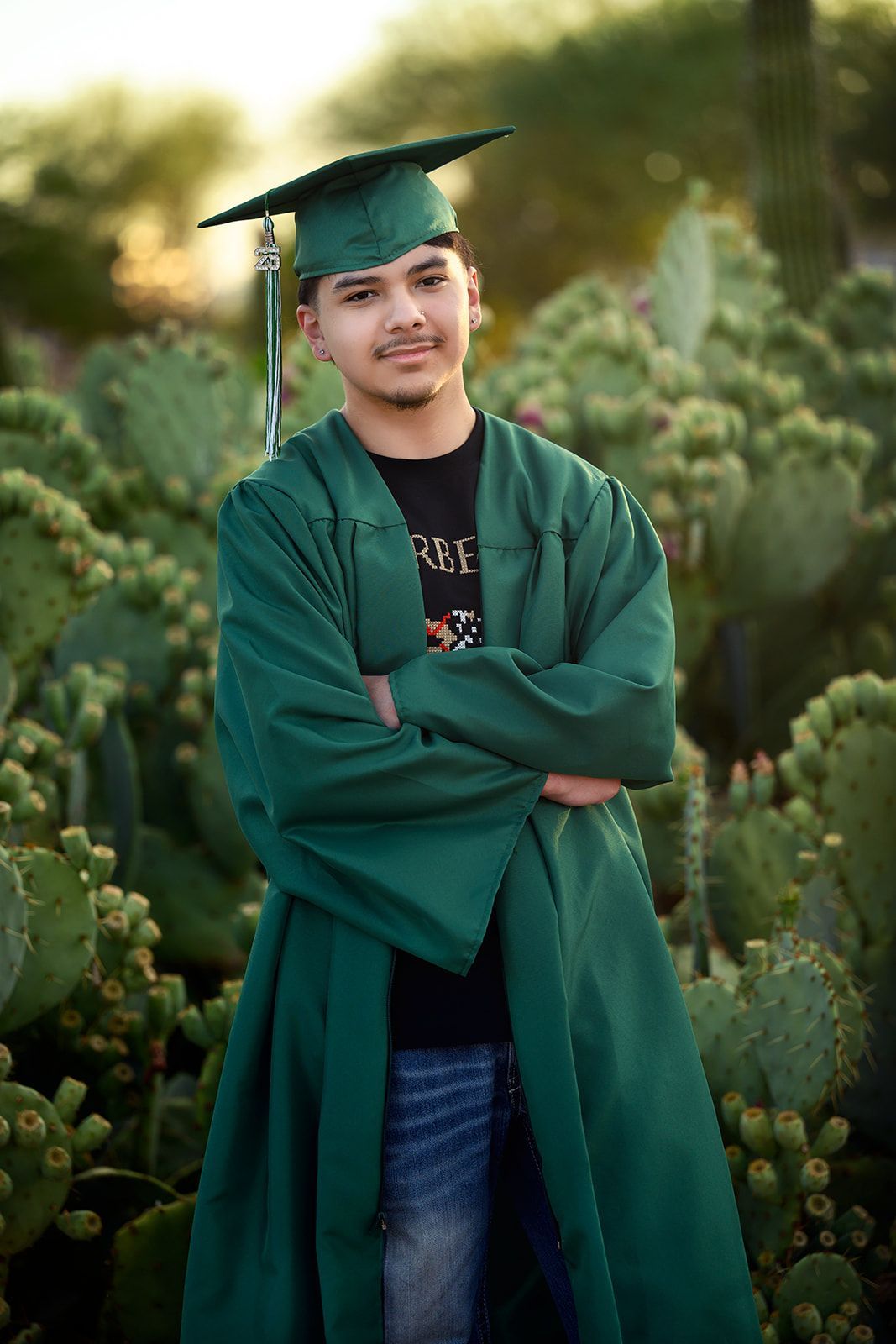 Person in a green graduation gown and cap, standing with arms crossed in a cactus garden.