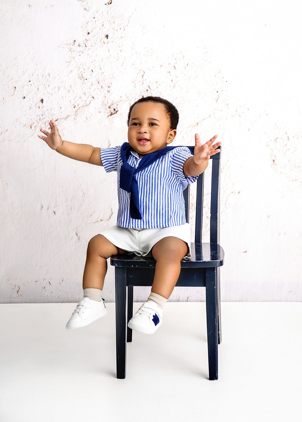 Baby in blue striped shirt sitting on a dark chair, arms outstretched against a white wall.