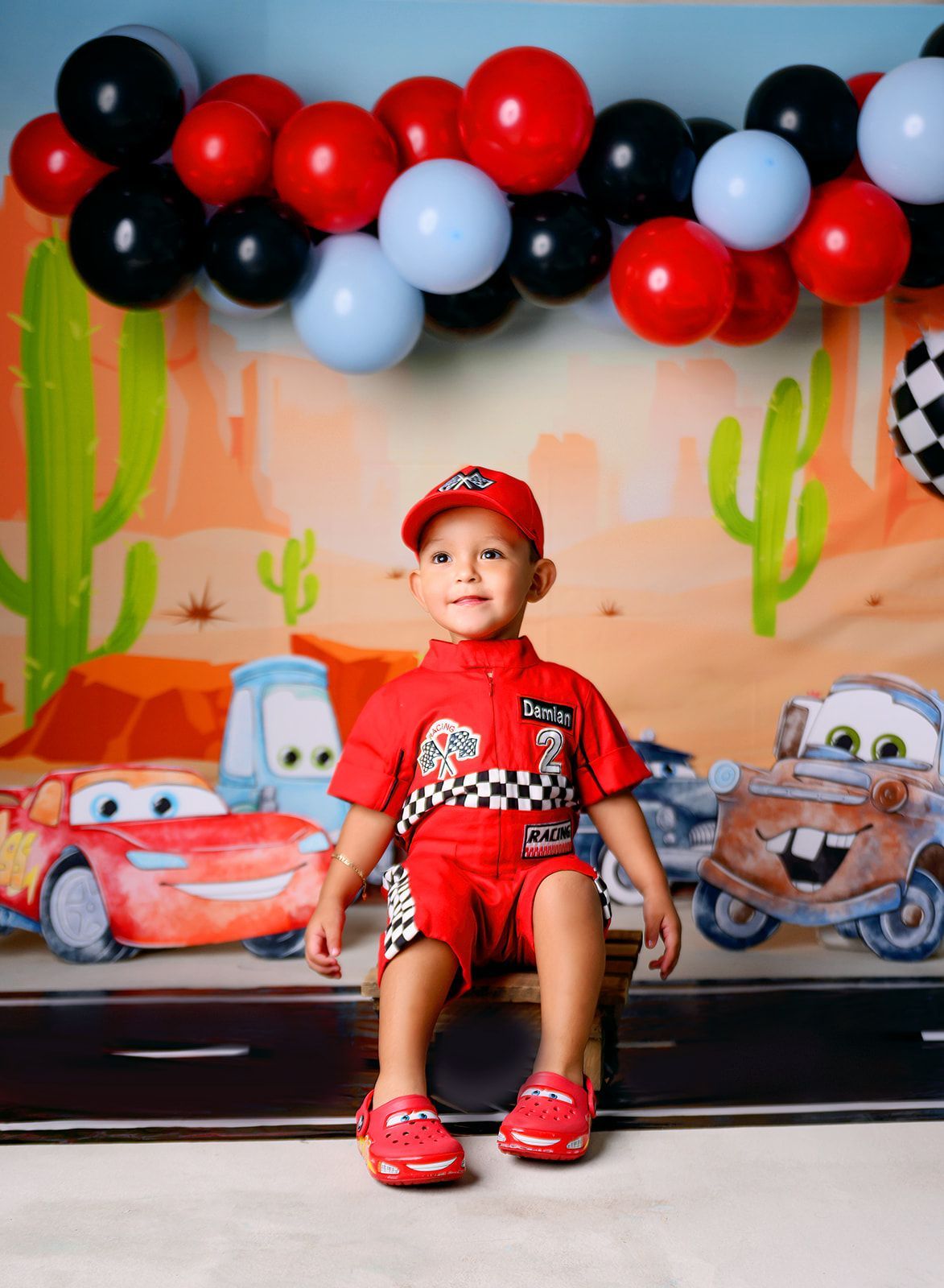 Child in red outfit and cap sitting before a cartoon race car backdrop with black, red, and white balloons.
