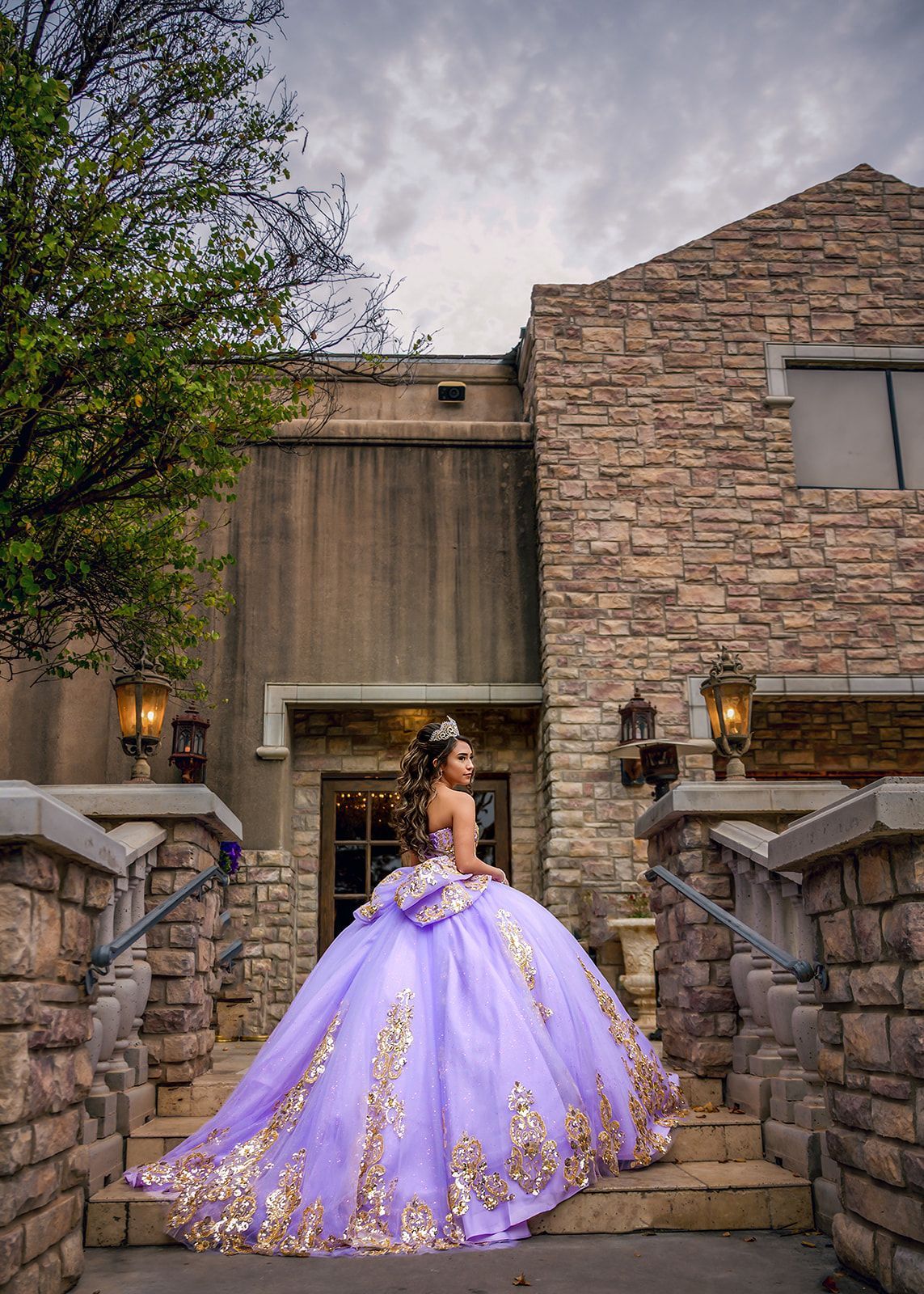 Woman in a lavender gown standing on stone steps between rustic buildings at dusk.