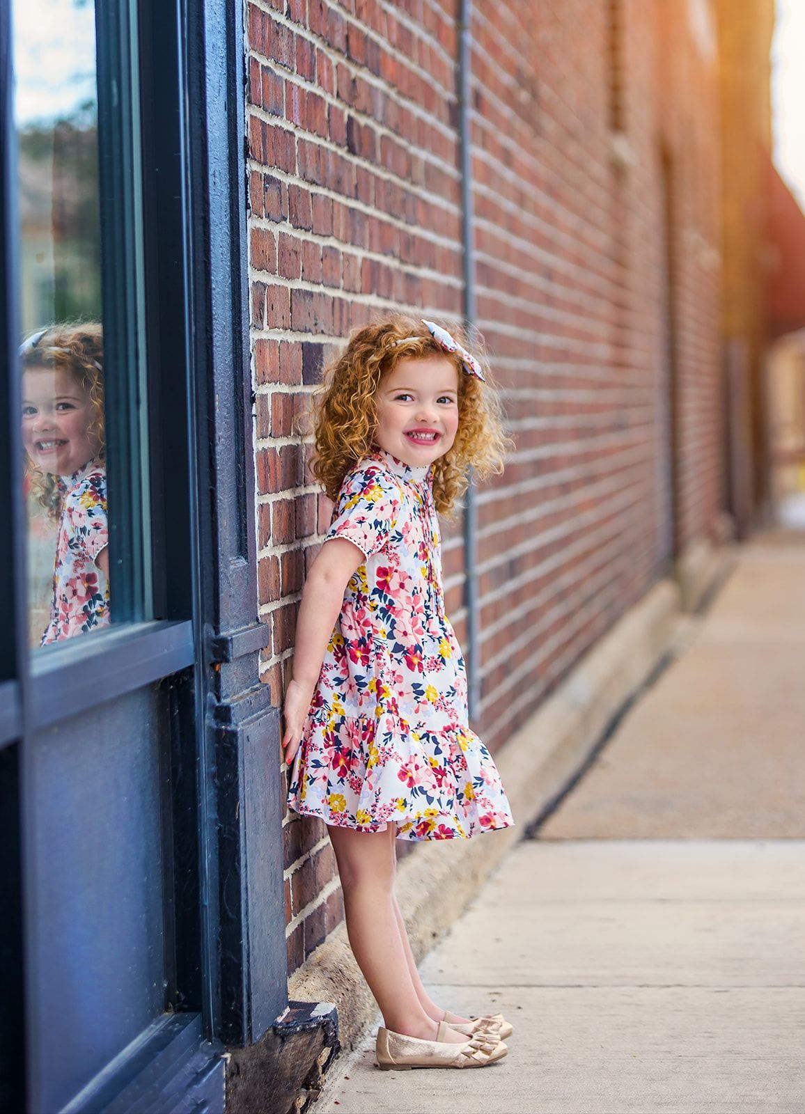 Child in a floral dress stands by a brick wall, smiling beside a window.