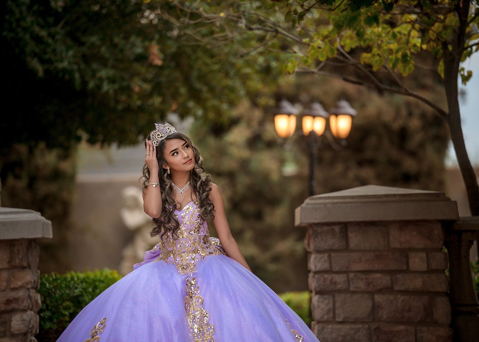 Woman in a lavender gown sitting on stone steps in a garden, looking up under warm lights.