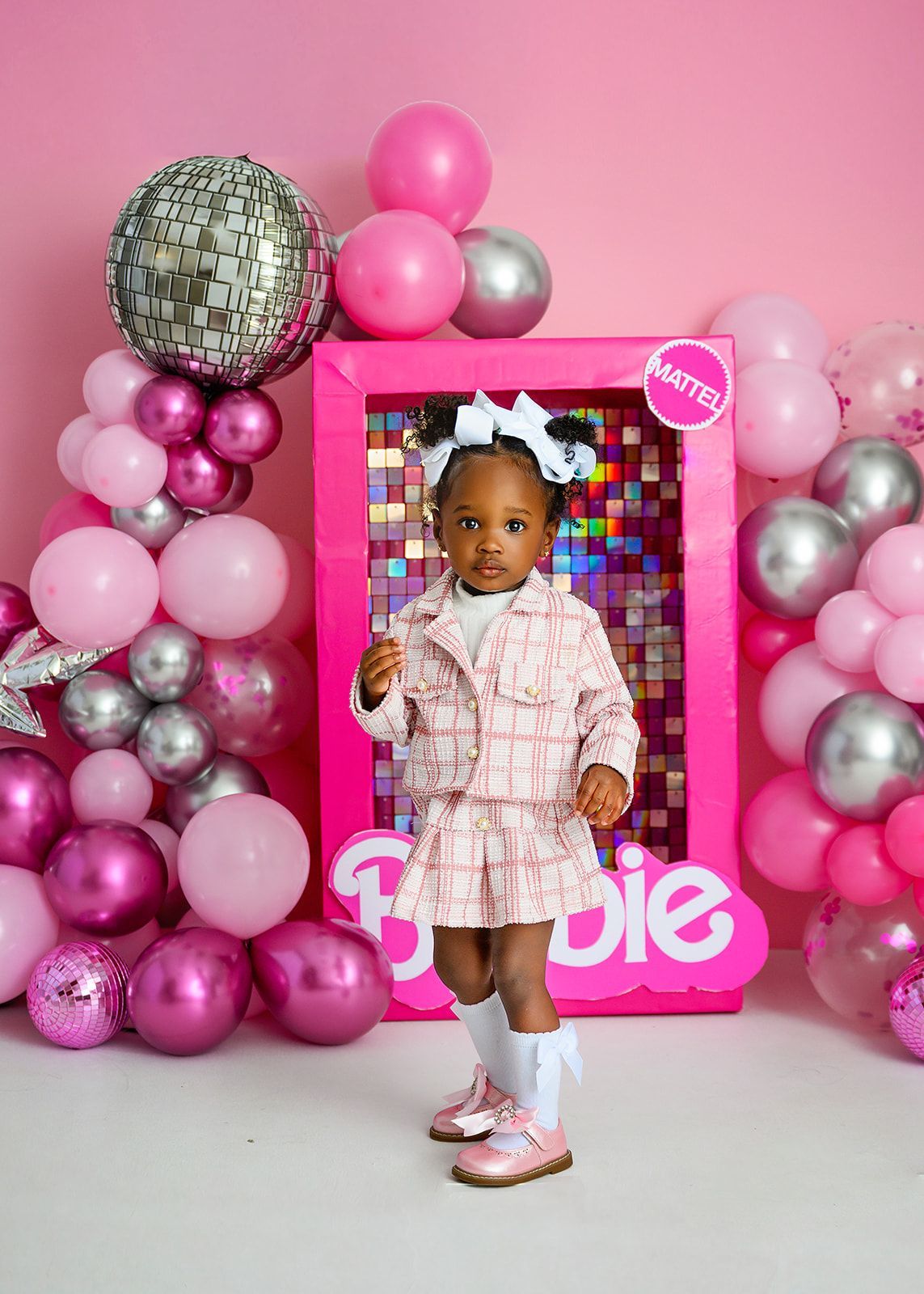 Child posing in pink outfit beside Barbie display and pink balloon decorations.