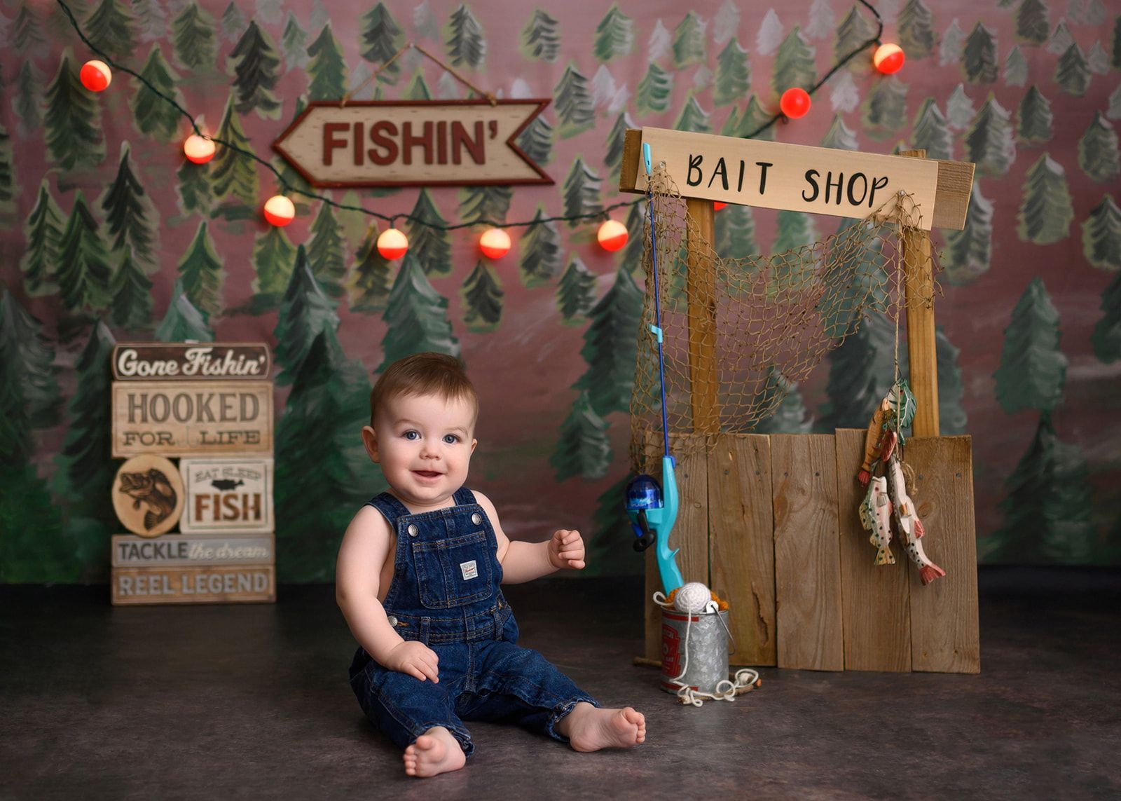 Baby sitting in a fishing-themed set with signs, string lights, and a bait shop backdrop.