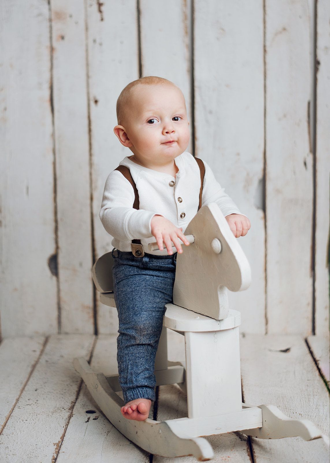 Toddler in white shirt and suspenders riding a white wooden toy horse against a rustic backdrop.