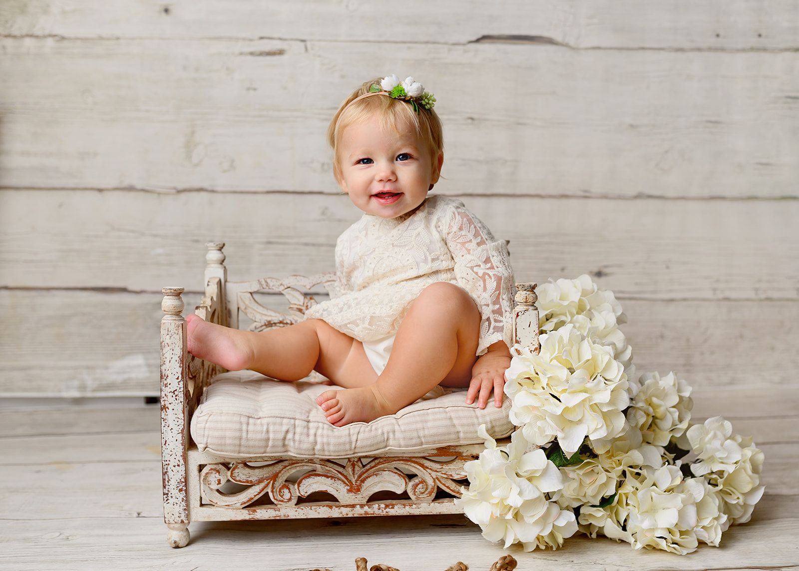 Smiling baby in a lace outfit sitting on a white wicker chair beside white flowers.