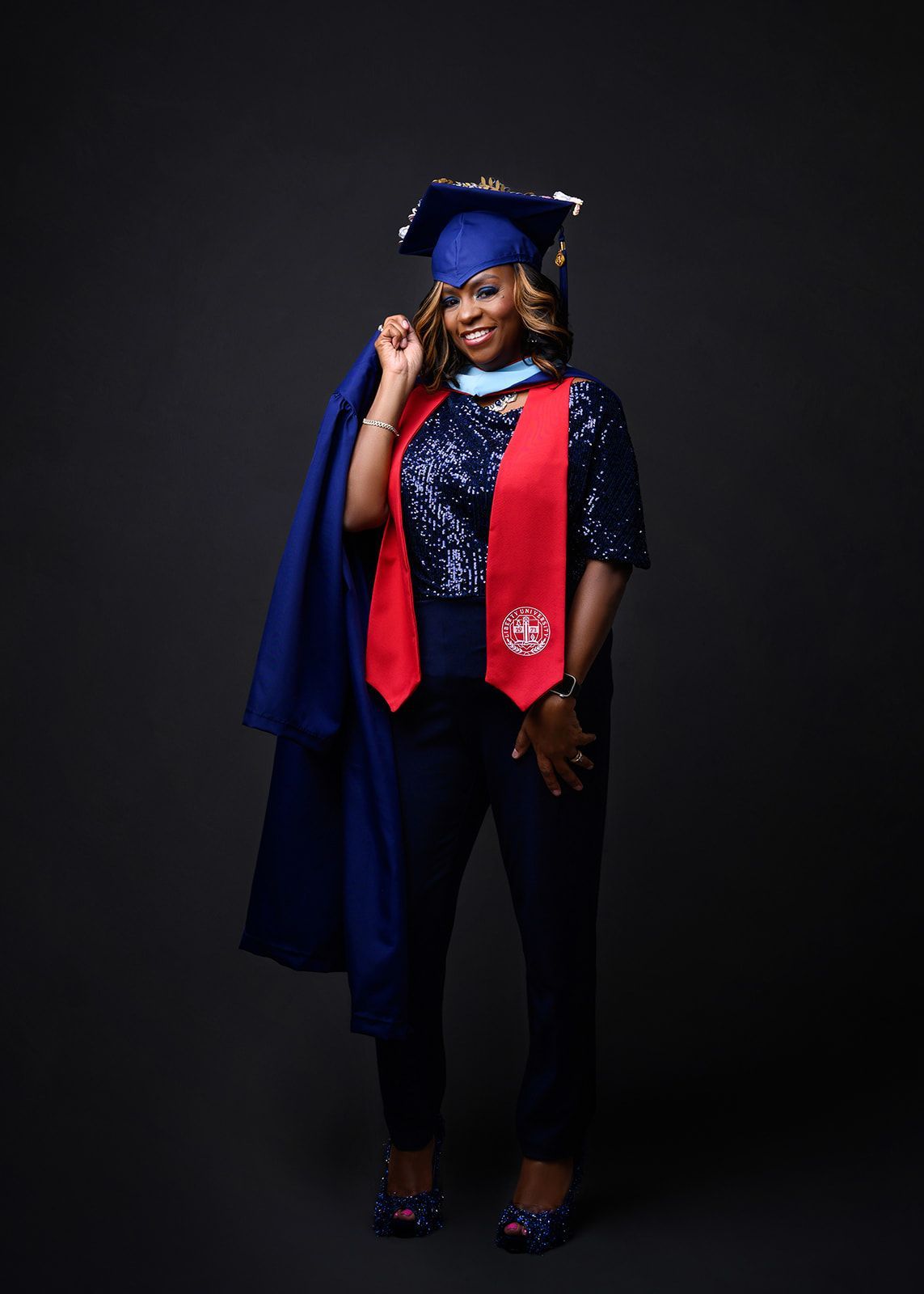 Graduate in blue cap and gown with red stole, standing and smiling against a dark background.