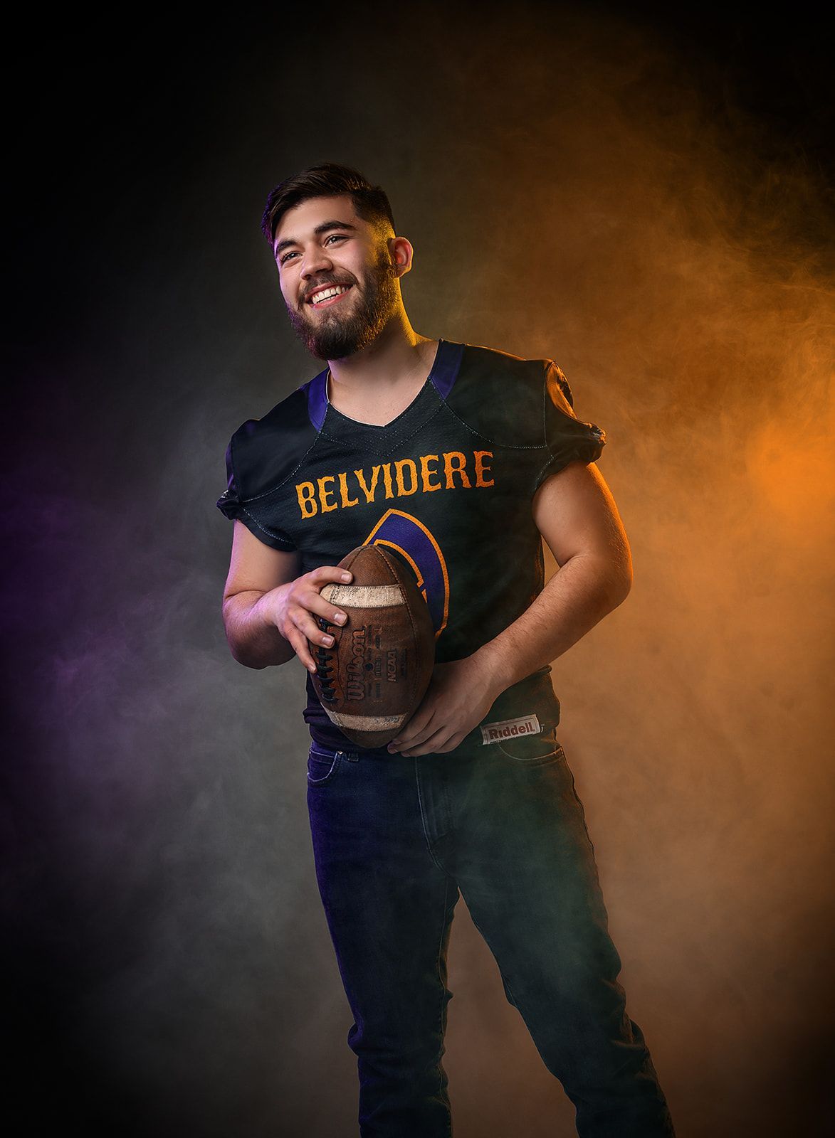 Smiling person in a BELVIDERE shirt holding a football against a dark, smoky background.