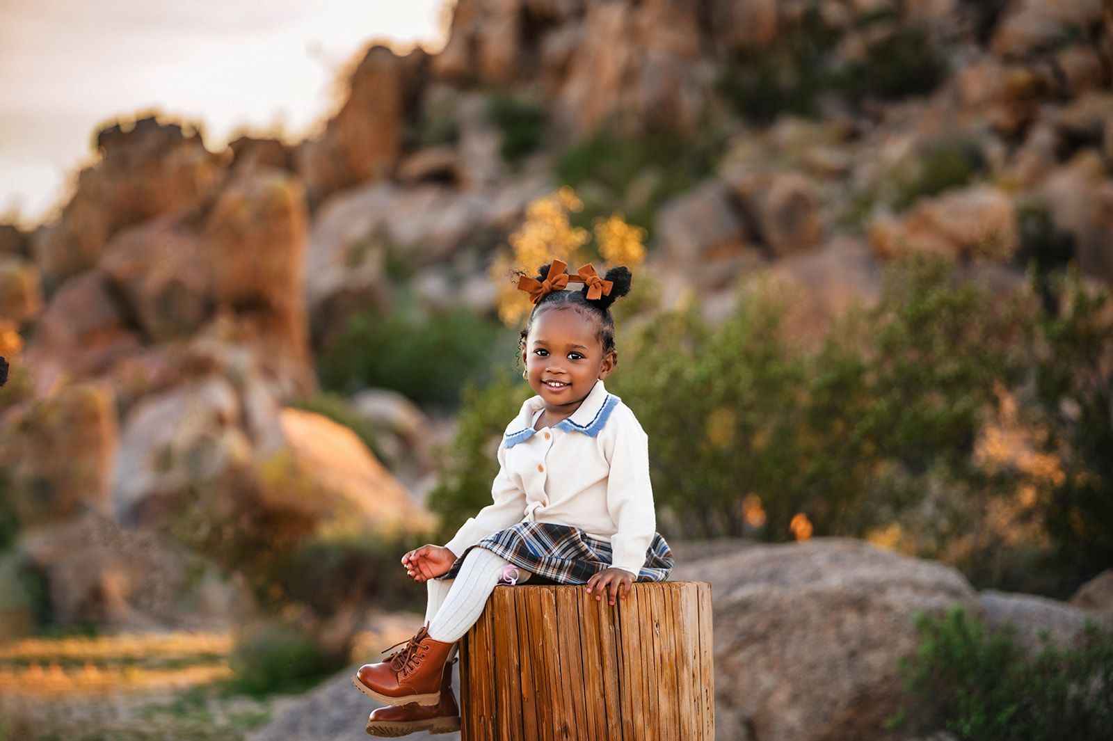 Toddler sitting on a tree stump outdoors, wearing a white outfit with braided hair and a bow.