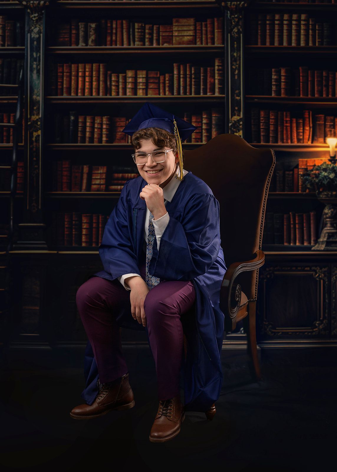 Person seated in a dark library, wearing a blue robe and cap, smiling with bookshelves behind them.