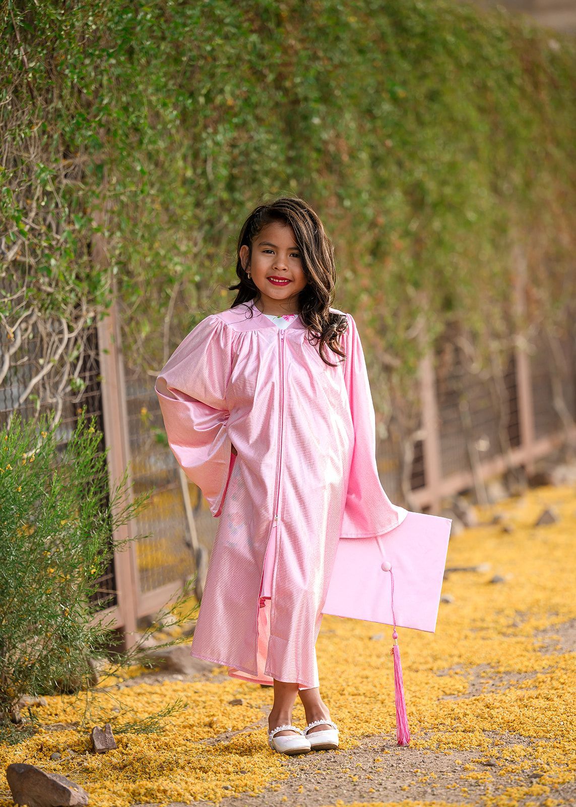 Young child in a pink dress and white shoes standing on a path covered with yellow petals by a green hedge.