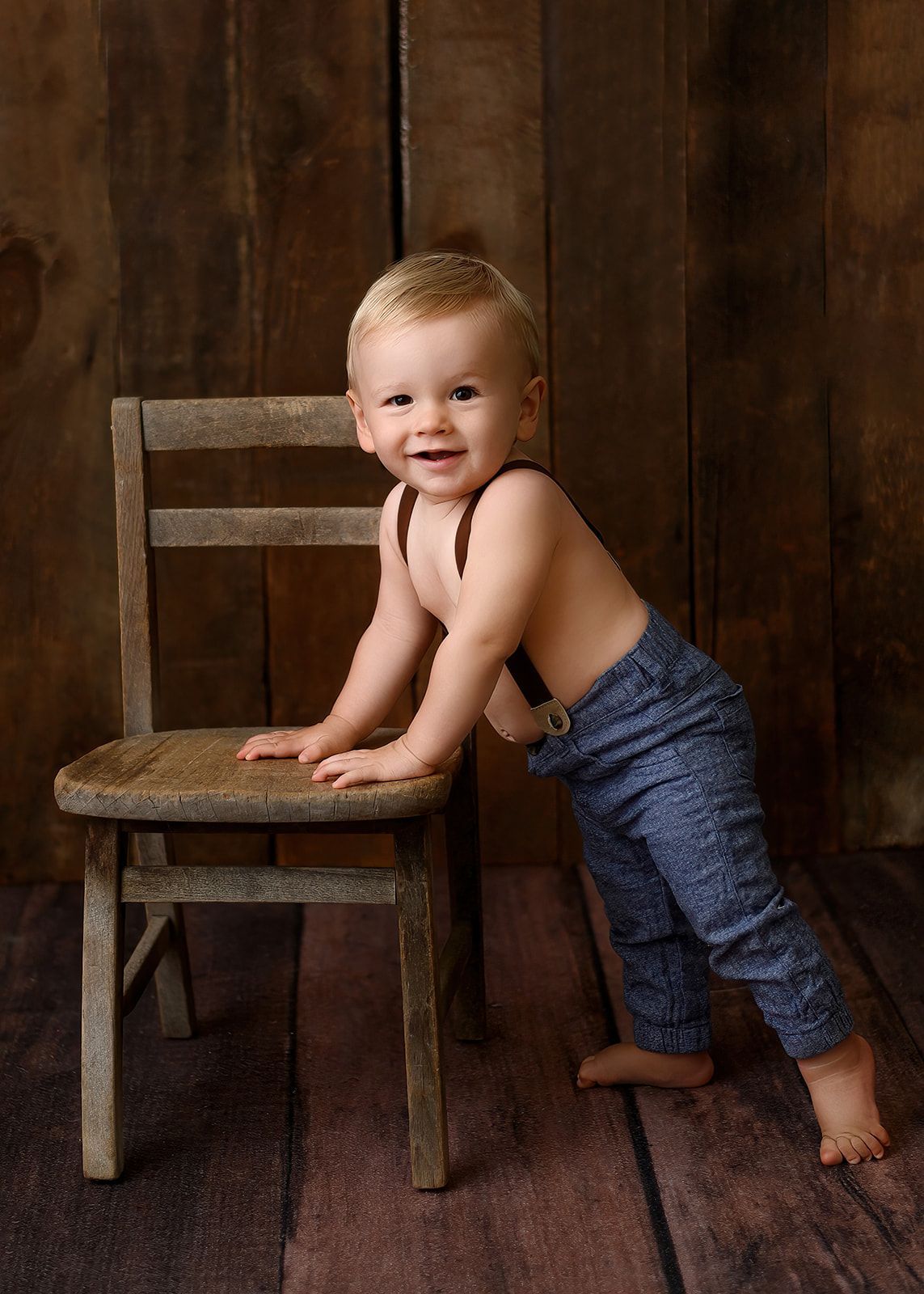 Smiling baby leaning on a wooden chair against a rustic wood backdrop.