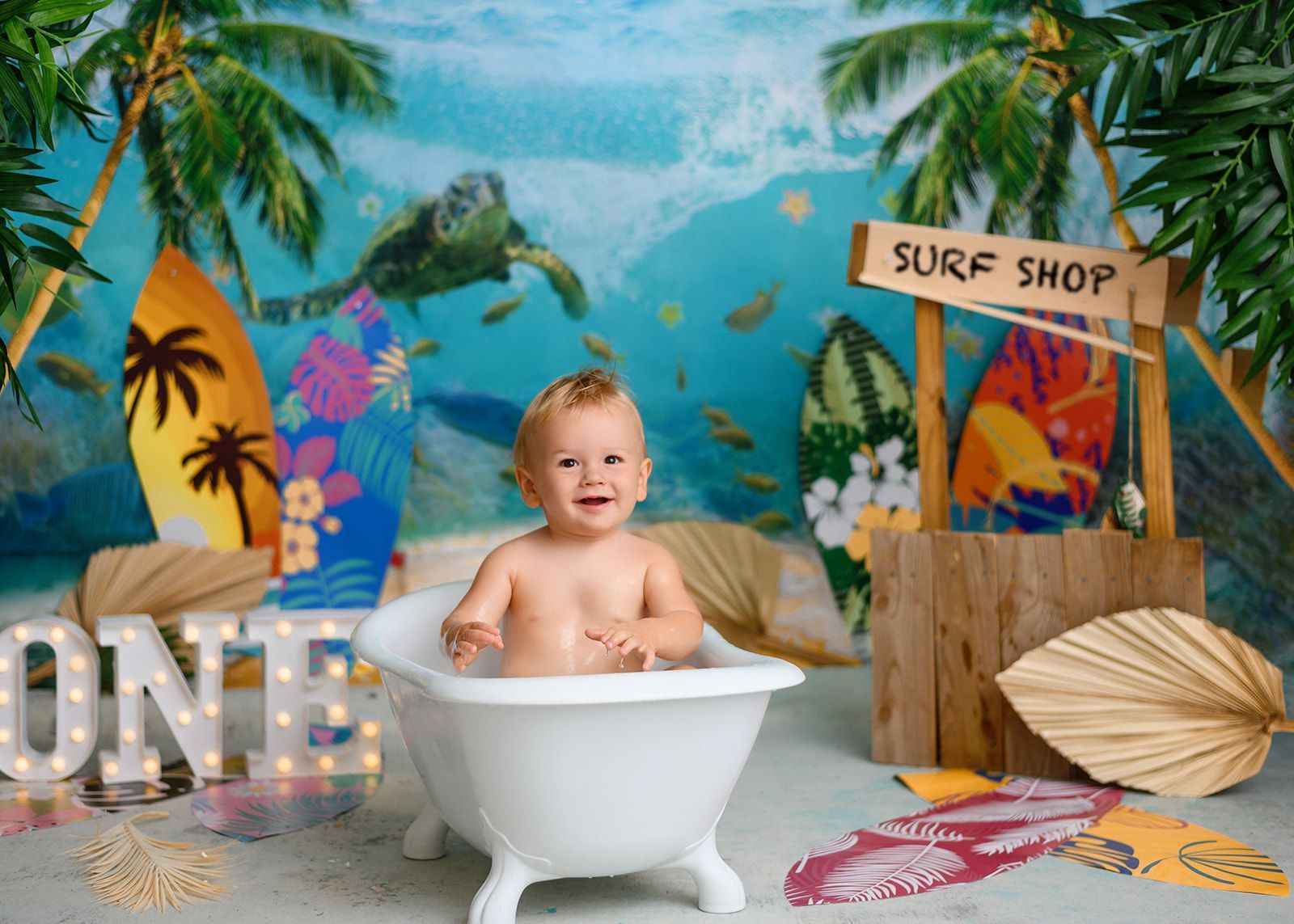 Baby smiling in a white tub at a colorful beach-themed surf shop backdrop.