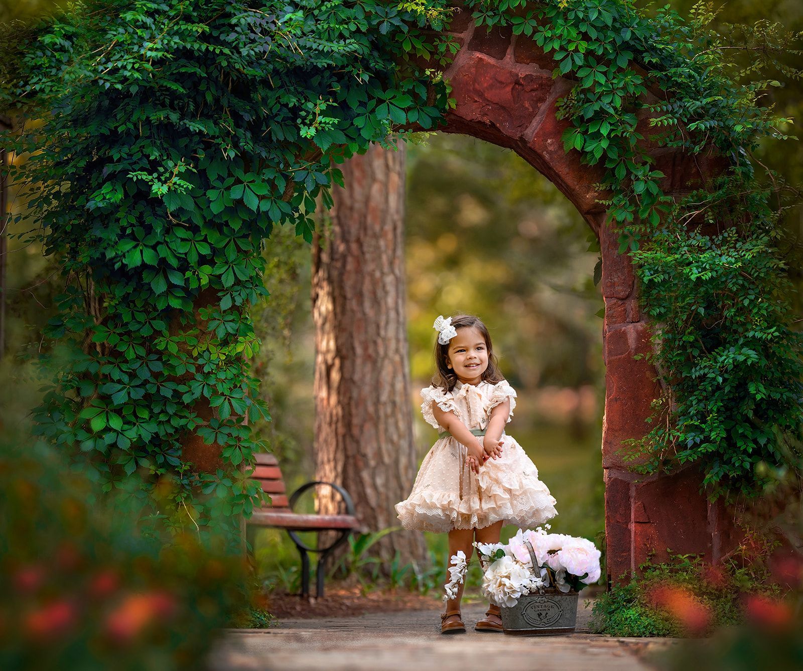 Young child in a white dress standing beneath a leafy garden arch beside a bench and flowers.