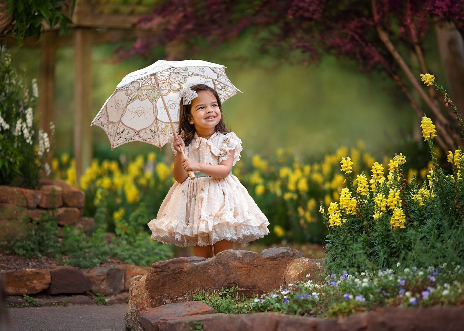 Child in a light dress holding a lace umbrella in a flower garden, standing on stone steps.