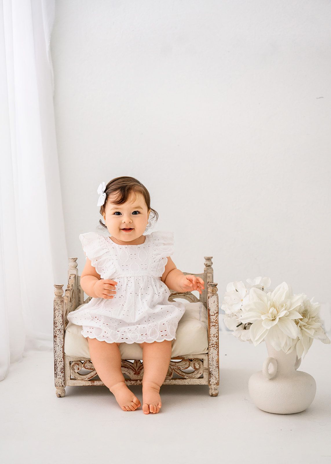 Baby in a white dress sitting on a tiny chair beside white flowers in a bright, minimalist room.