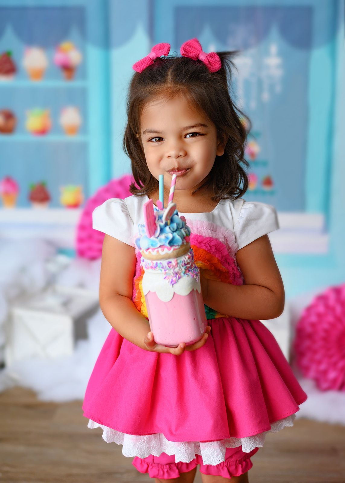 Young child in a pink dress holding a colorful milkshake, standing indoors with a bright pastel backdrop.