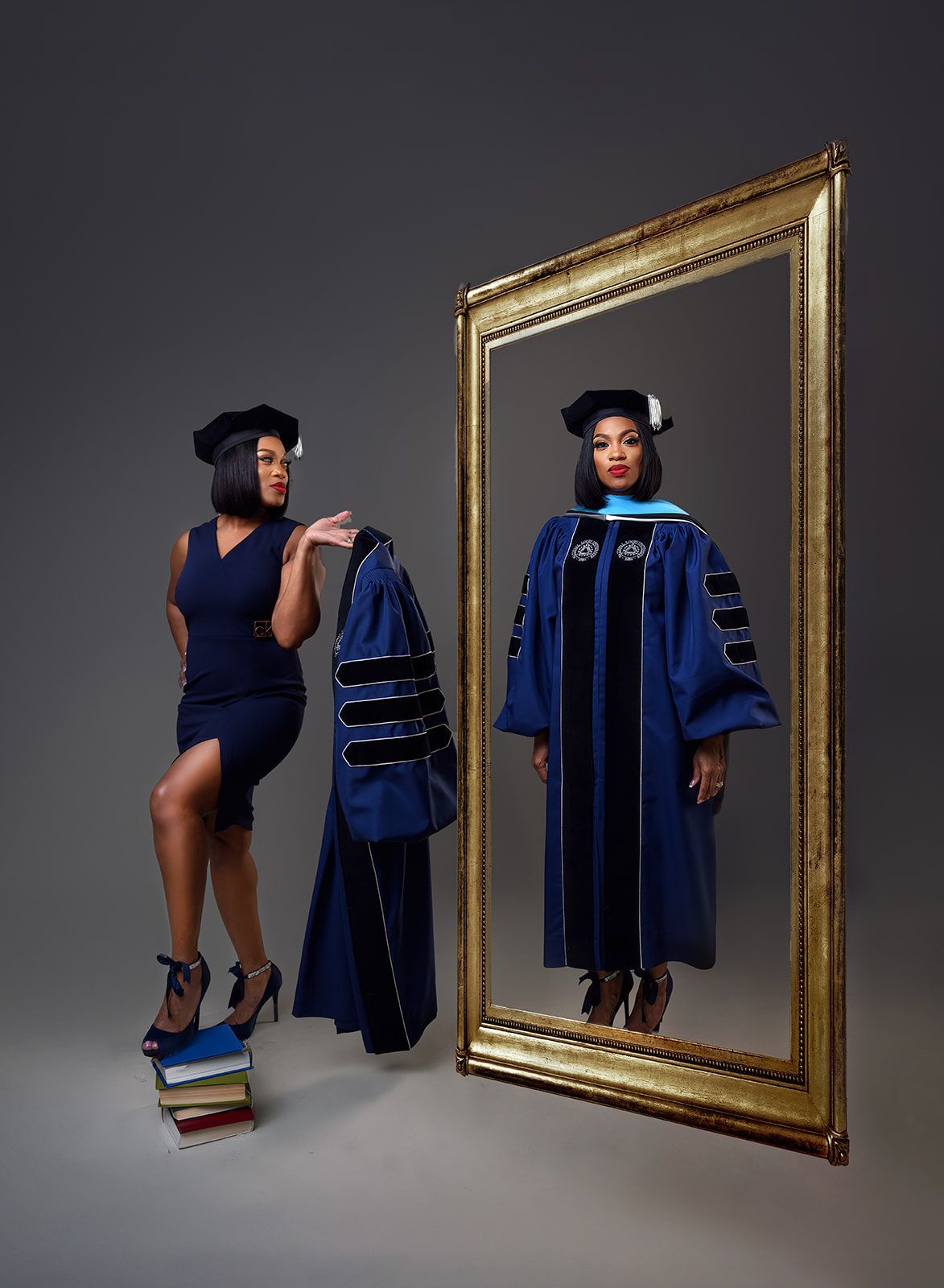Woman in blue graduation gown beside mirror and stacked books in a studio setting.