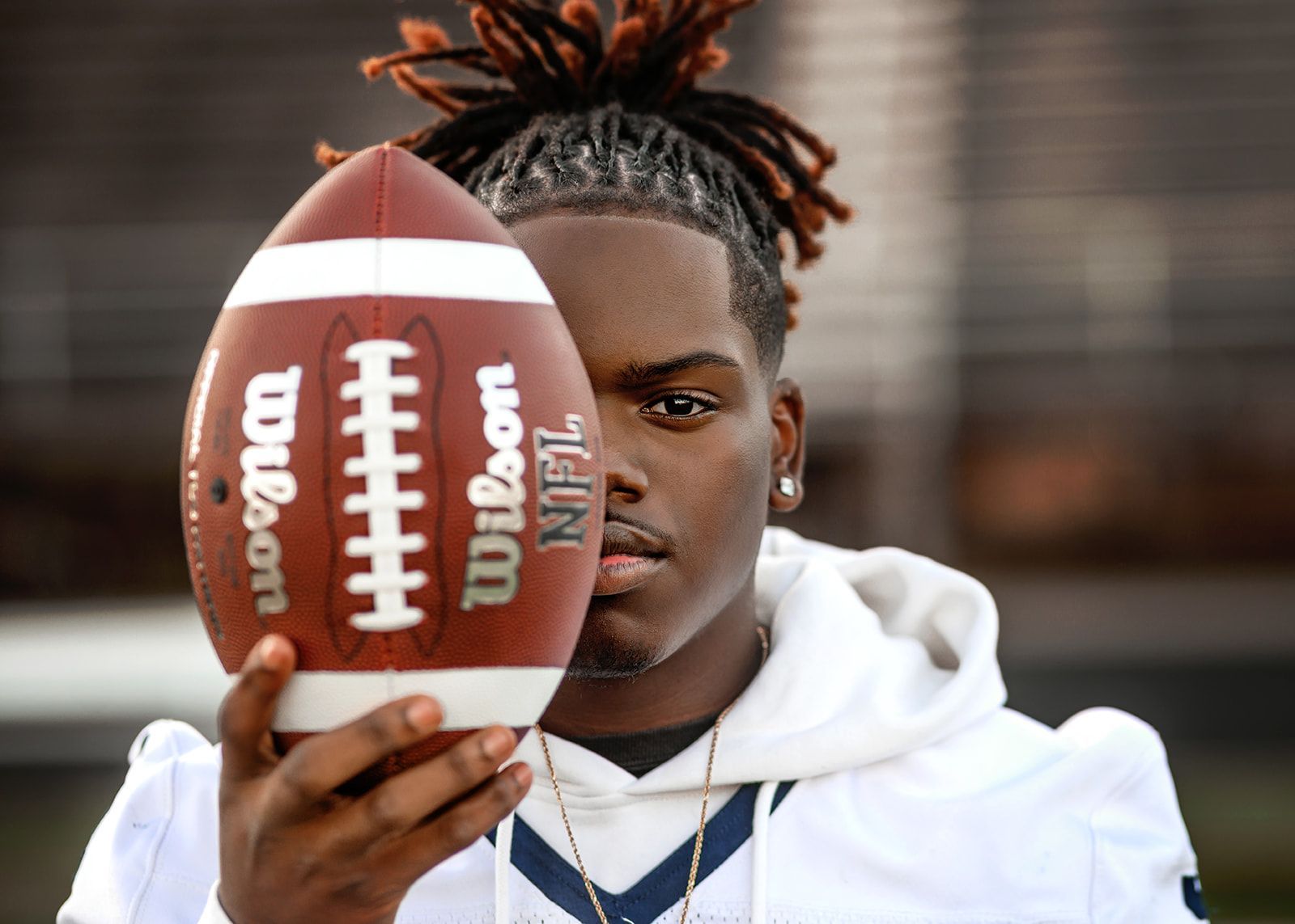 Football player holding a football in front of his face, wearing a white hoodie.