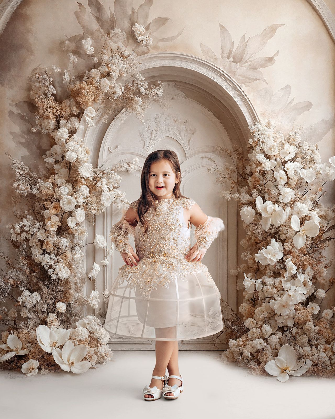 Child standing in a cream floral dress against a beige floral arch backdrop.