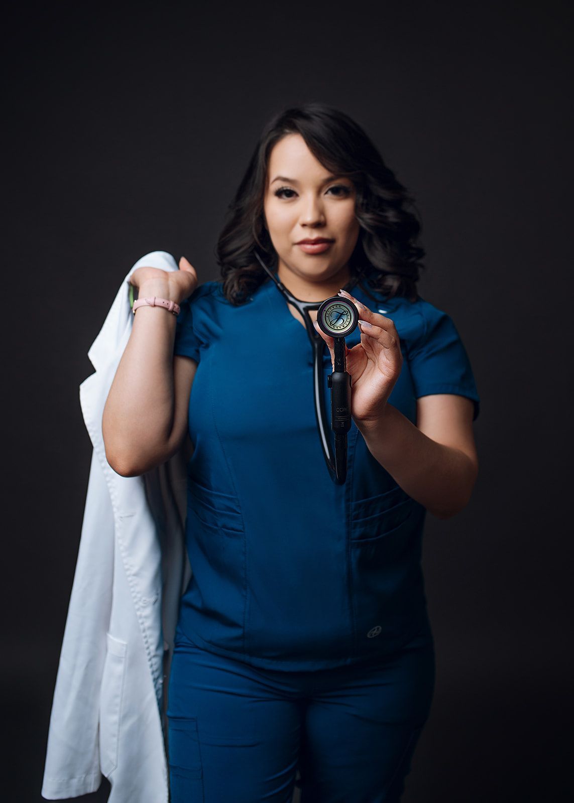 Woman in blue scrubs holding a white coat and a stethoscope against a dark background.