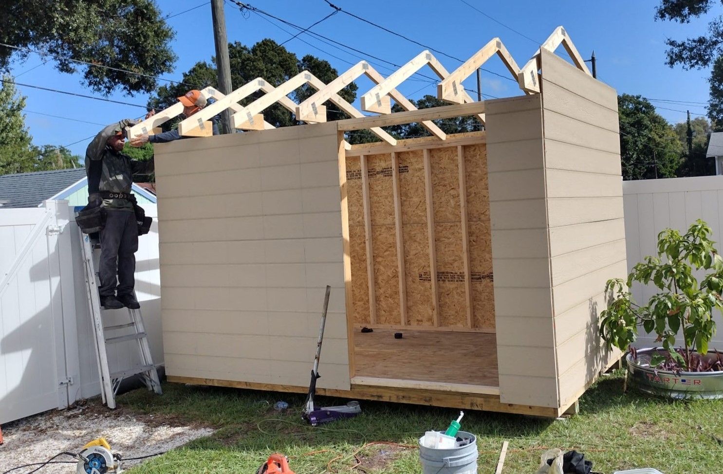 A man is standing on a ladder working on the roof of a shed.
